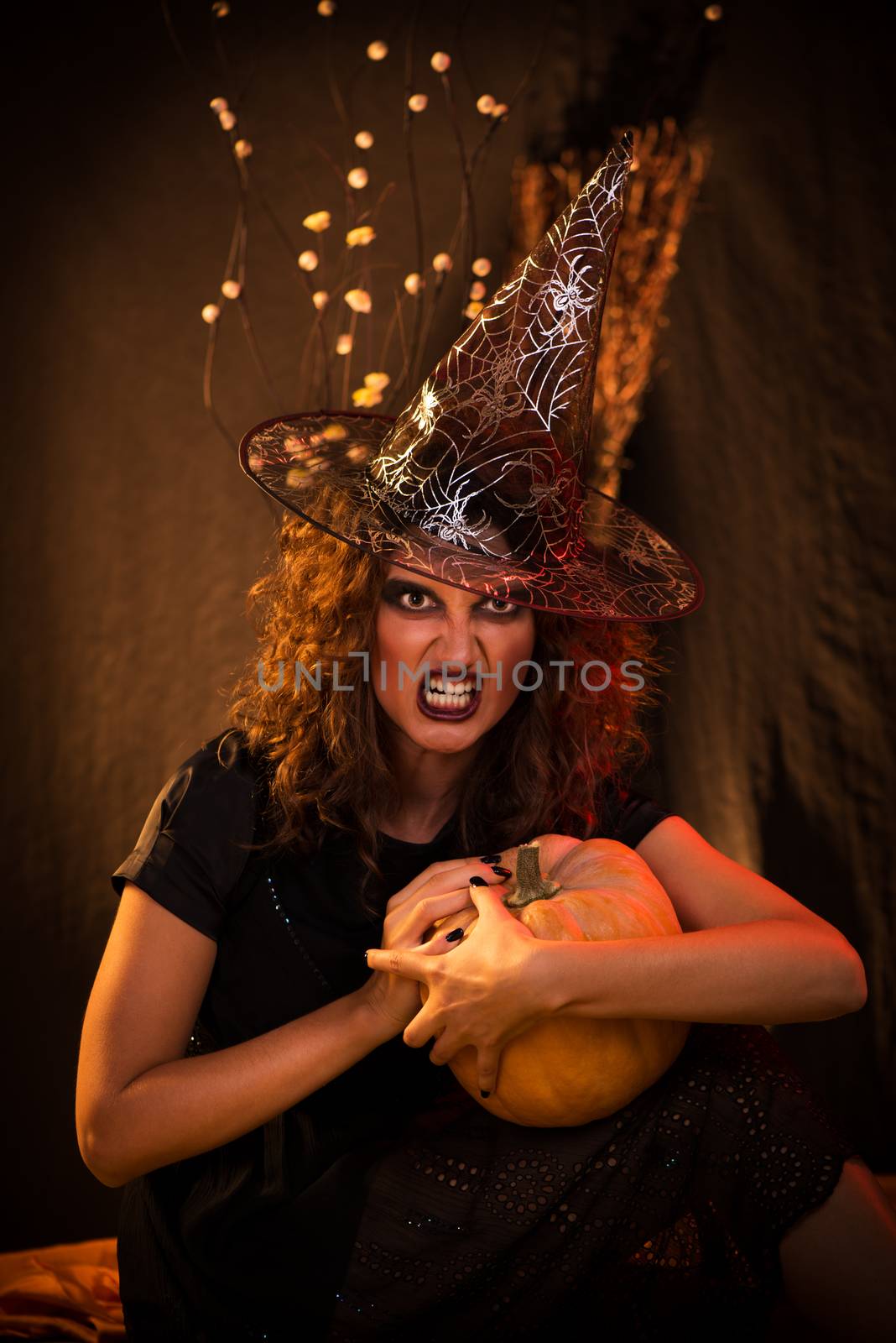 Young woman with evil face dressed like a witch. She wears dark clothing and holding a pumpkin in hands.