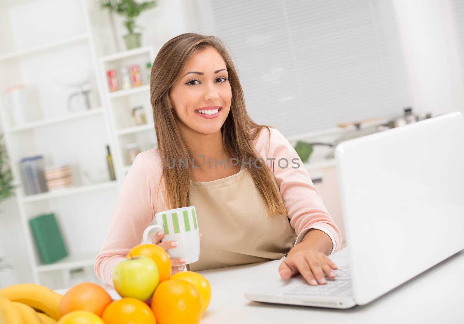 Beautiful young woman in the kitchen drinking coffee and reading recipe on laptop. Looking at camera.
