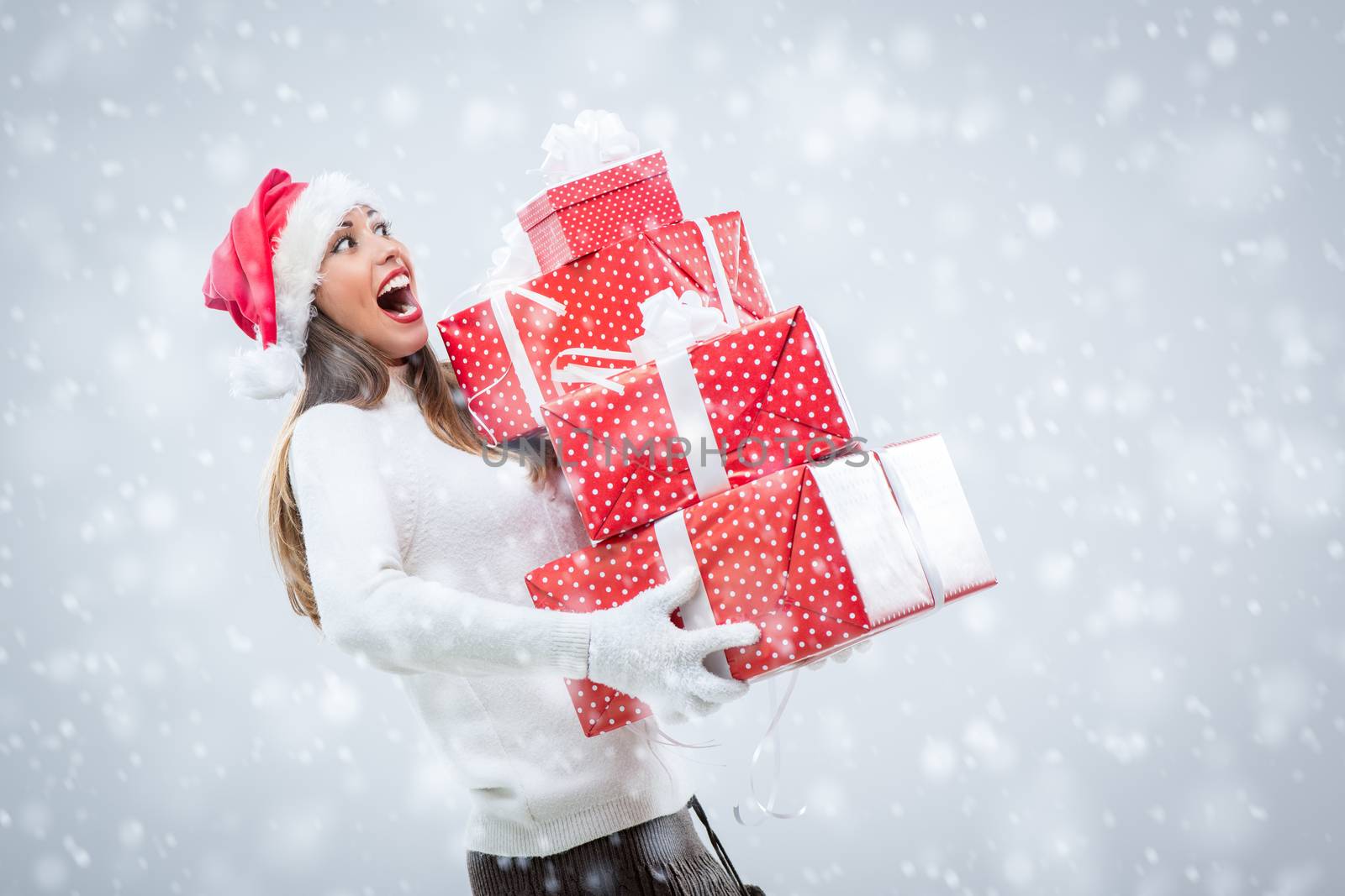 Cheerful beautiful young woman wearing santa hat and holding many Christmas gifts while snowing.