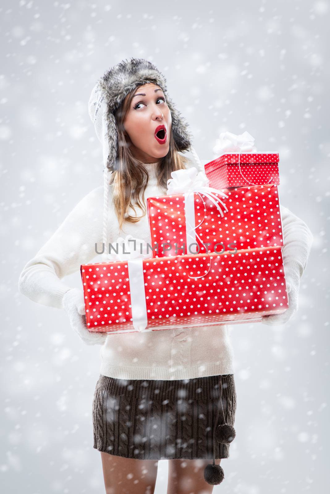 Cheerful beautiful young woman in warm clothing holding many Christmas gifts while snowing.