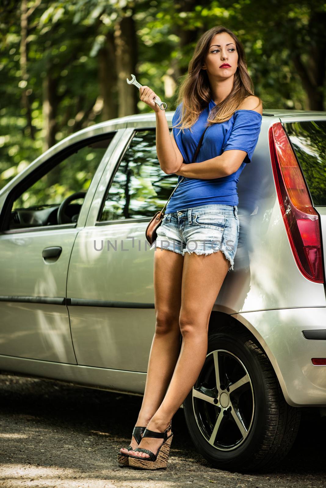 Beautiful young woman with a silver car that broke down on the road in forest. She has standing by a car and holding wrench.