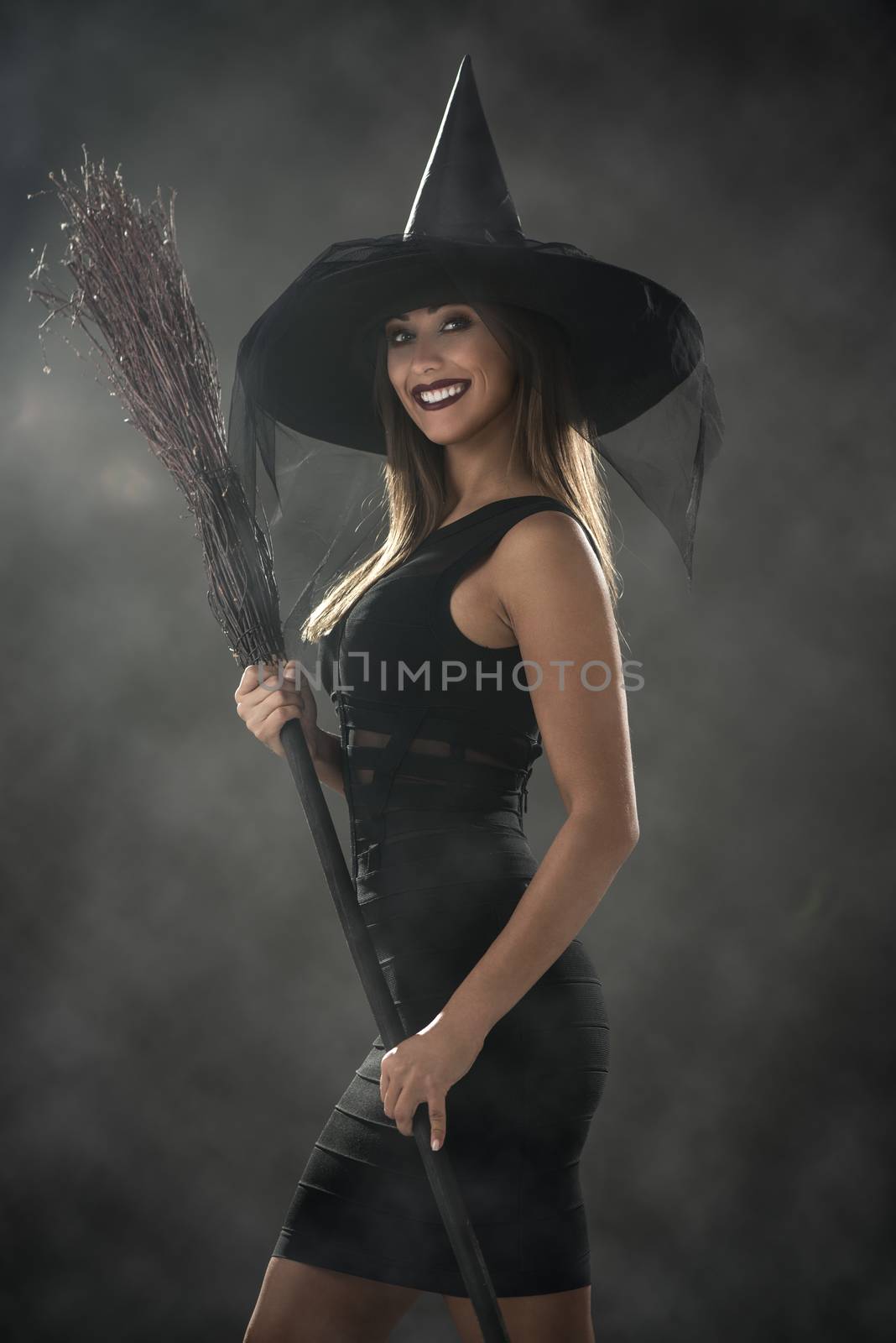 Young woman dressed like a witch. She is in dark clothing and holding a broom.
