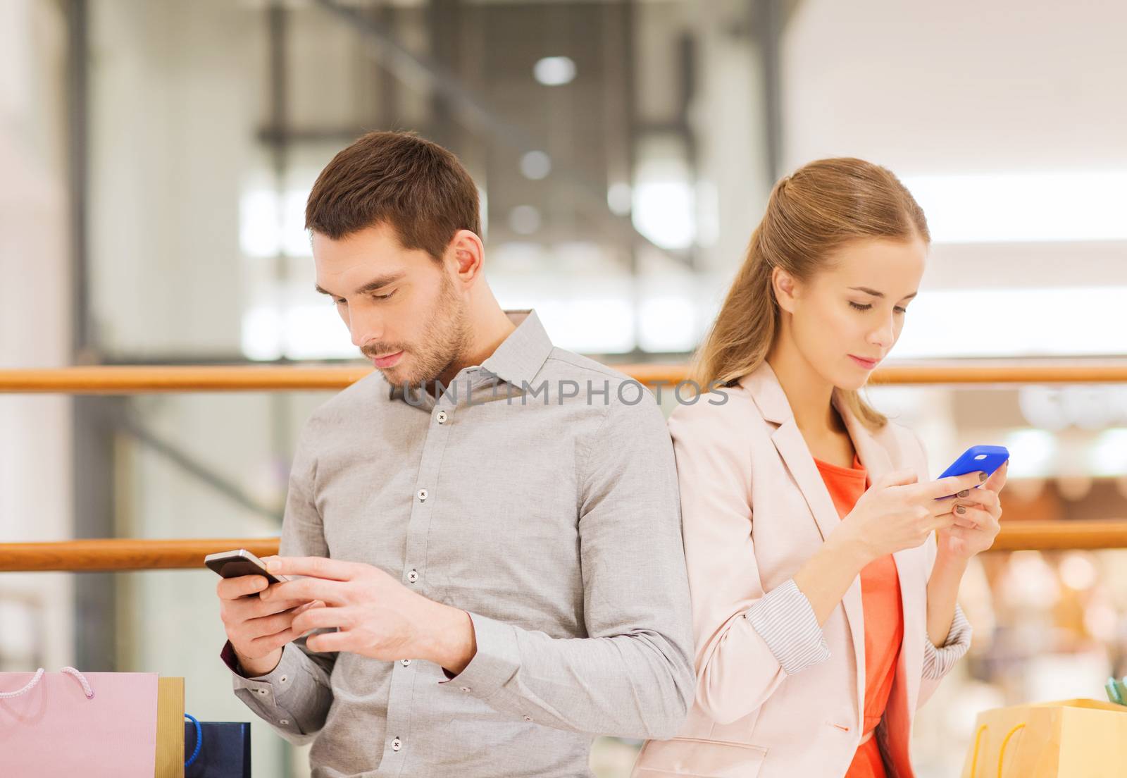 sale, consumerism, technology and people concept - young couple with shopping bags and smartphones in mall