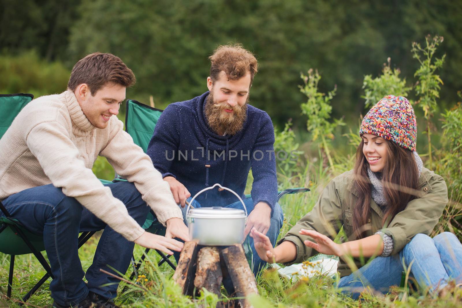 adventure, travel, tourism and people concept - group of smiling friends cooking food in dixie and warming hands sitting around bonfire outdoors