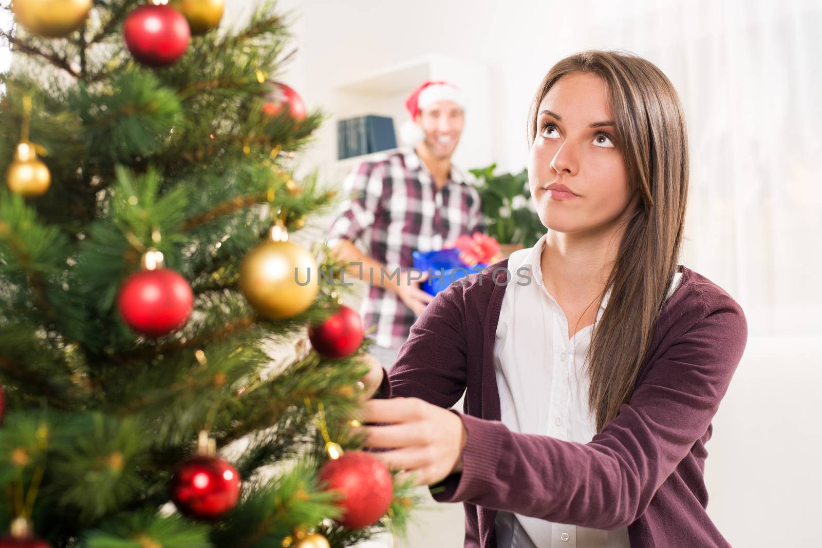 Young beautiful girl decorates a Christmas tree while her boyfriend holds a Christmas gift and he wants to surprise her.
