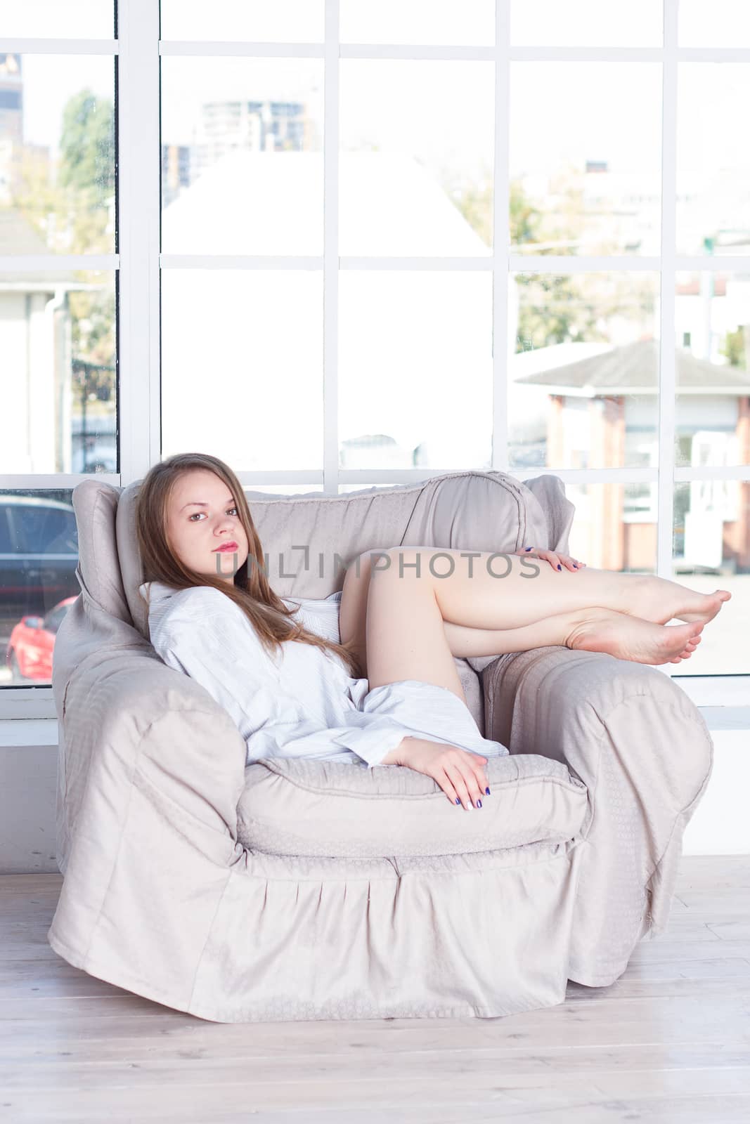 Young woman at home sitting on modern chair in front of window relaxing in her living room