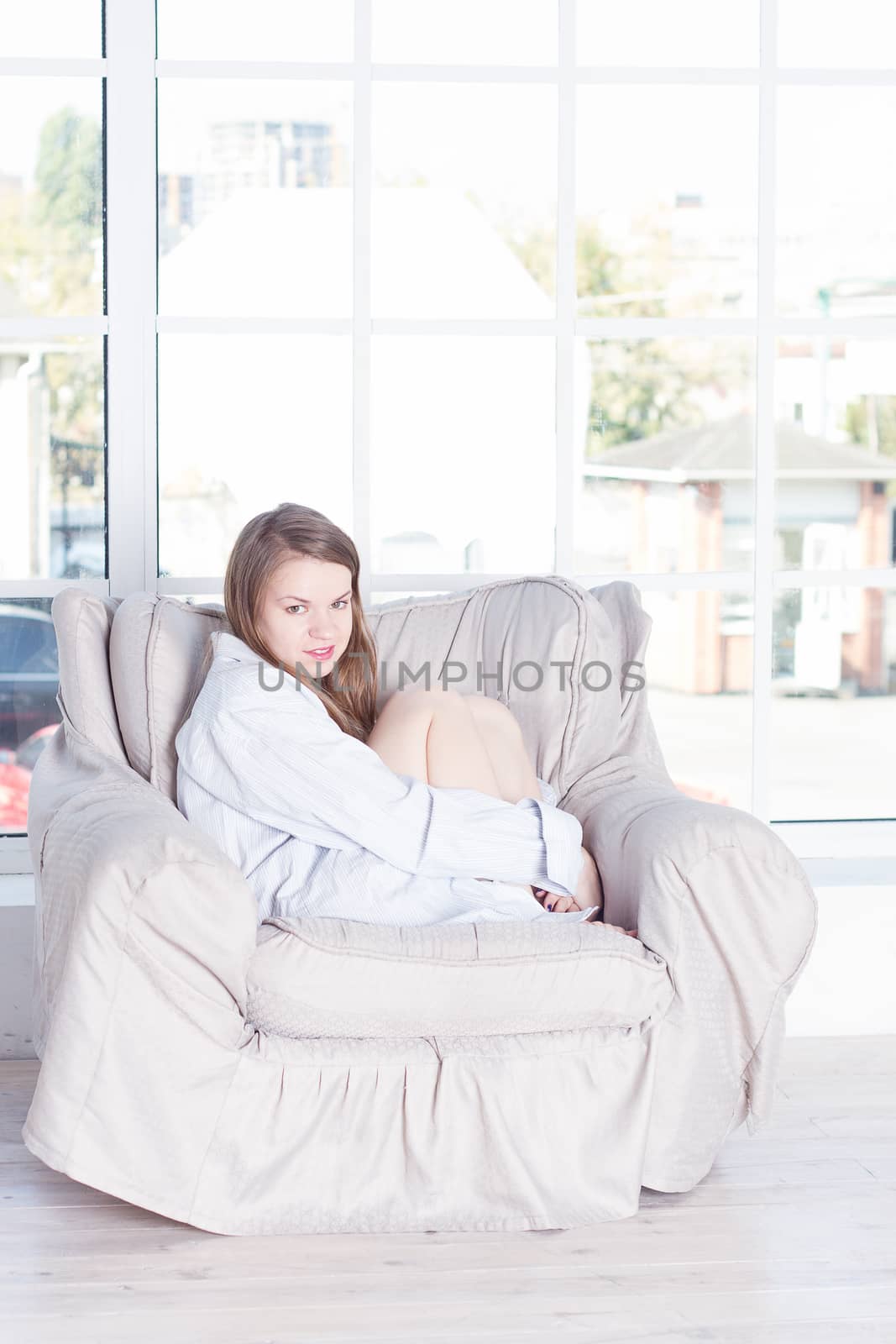 Young woman at home sitting on modern chair in front of window relaxing in her living room