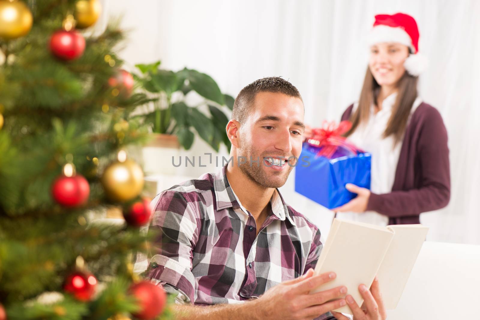 Young beautiful man reading book while his girlfriend holds a Christmas gift and she wants to surprise him.