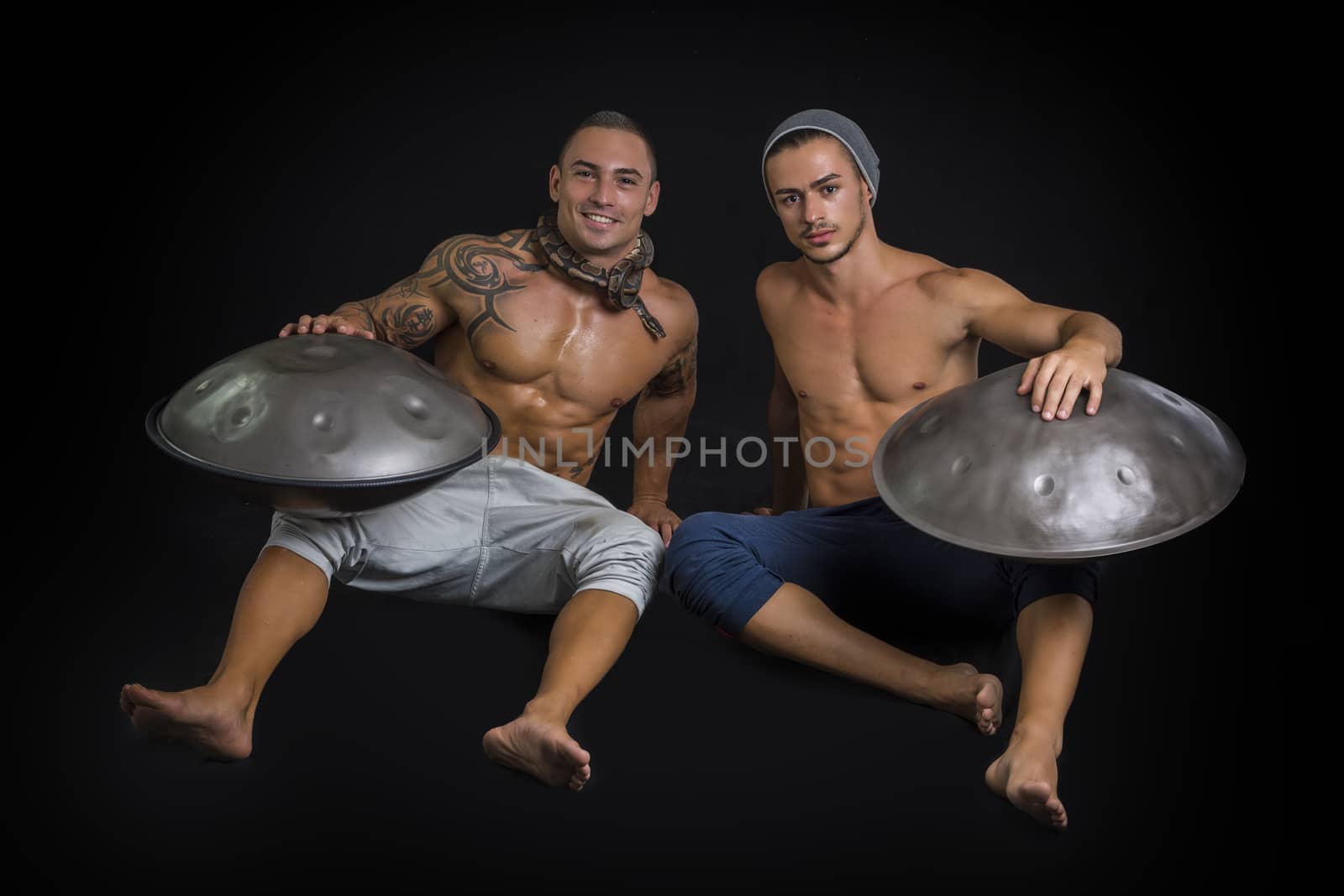 Two Exotic Male Drummers Drumming with Hands on Steel Pan Drums While Seated Beside Each Other in Studio, Isolated on Black Background