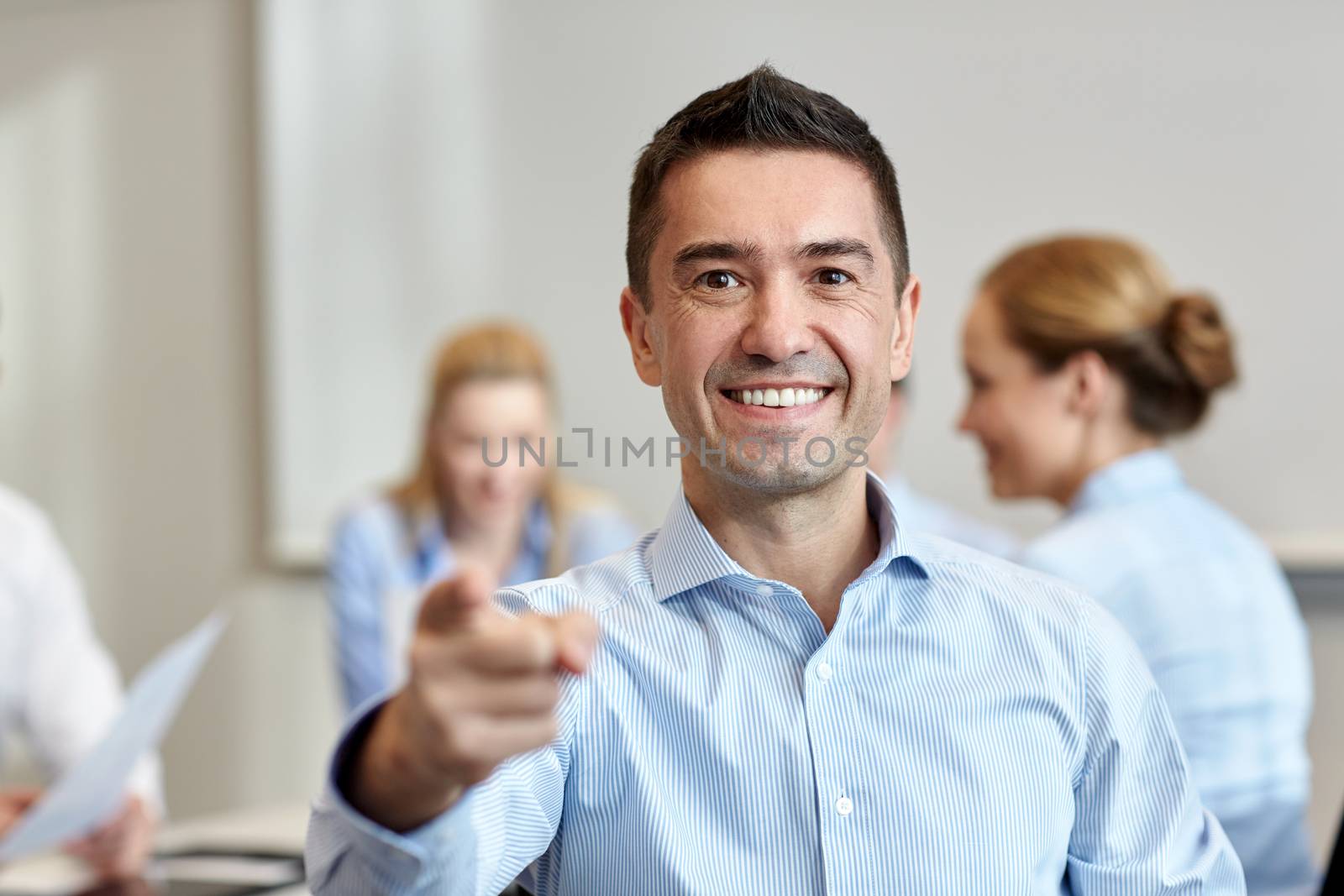 business, people, gesture and teamwork concept - smiling businessman pointing finger to you with group of businesspeople meeting in office