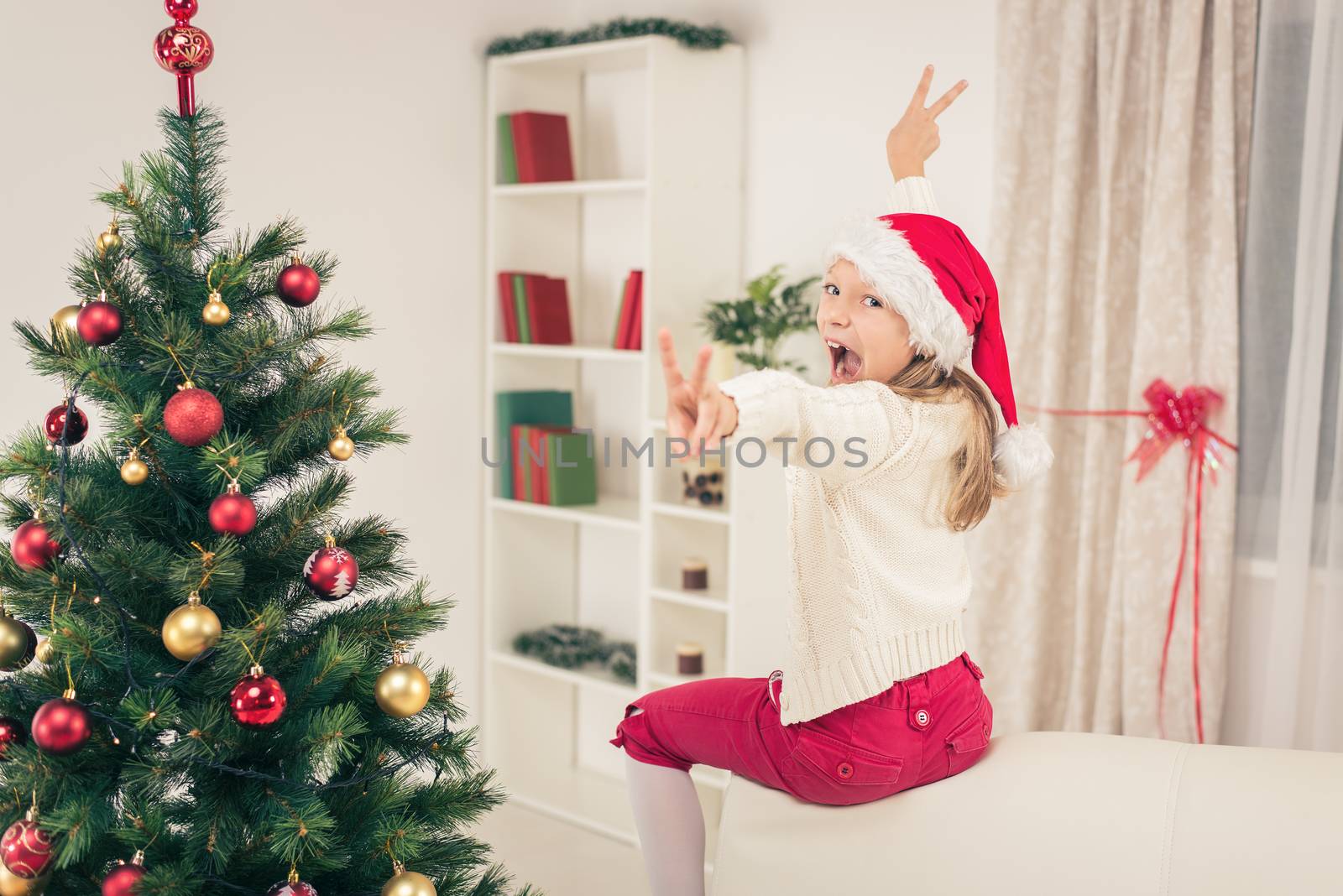 Cheerful little girl wearing Santa's hat and having fun at home at Christmas Time. Lookin at camera.