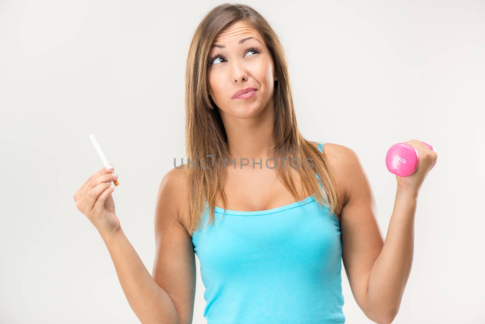 Beautiful young woman holding cigarette in one hand and dumbbell in second hand and thinking.