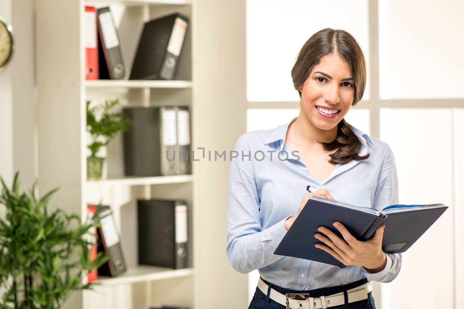Beautiful young smiling businesswoman standing in office and holding planner. In the background you can see the shelves with binders. Looking at camera.