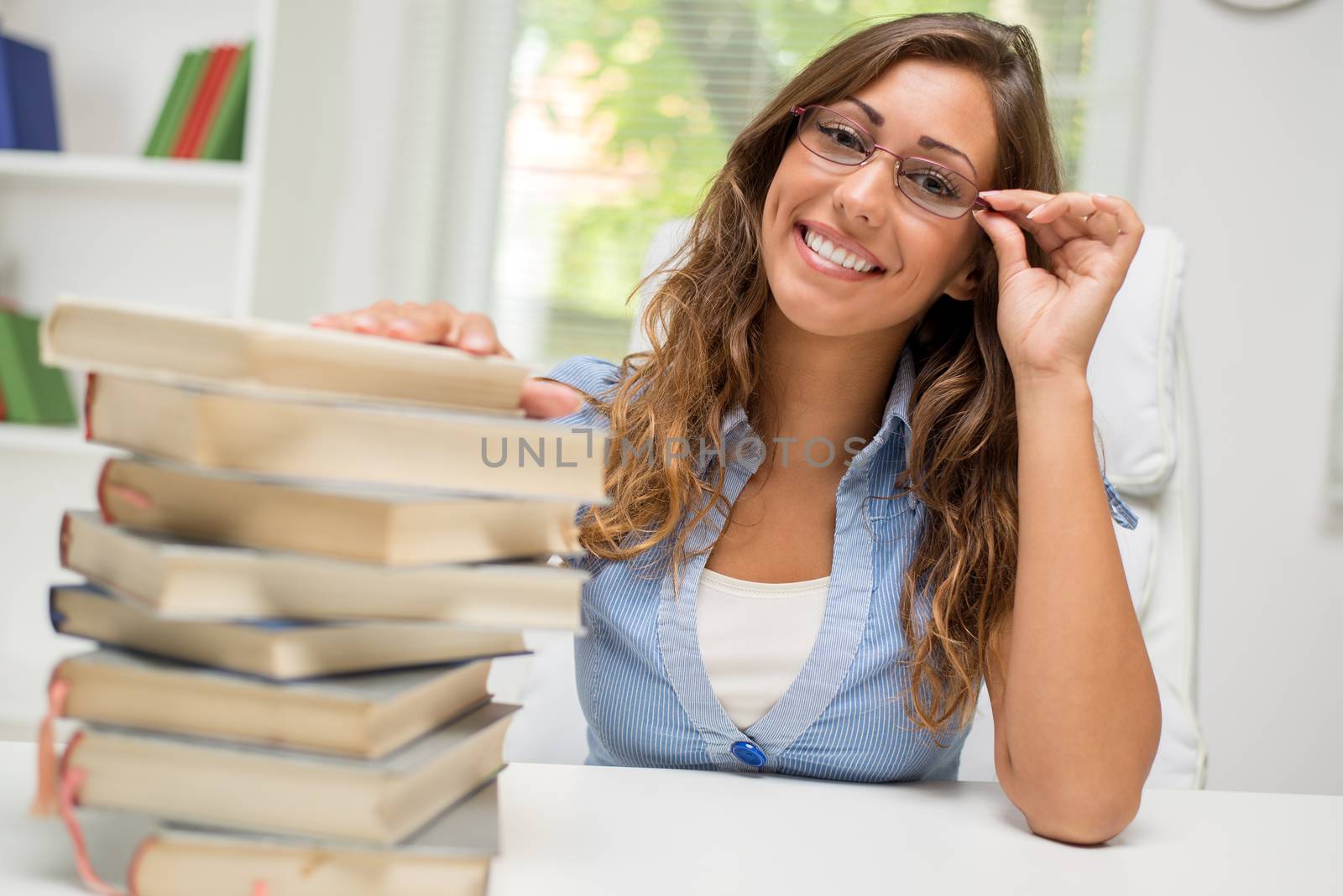 Beautiful teenage girl with eyeglasses sitting in library and holding hand on stack of books. Looking at camera and smiling.