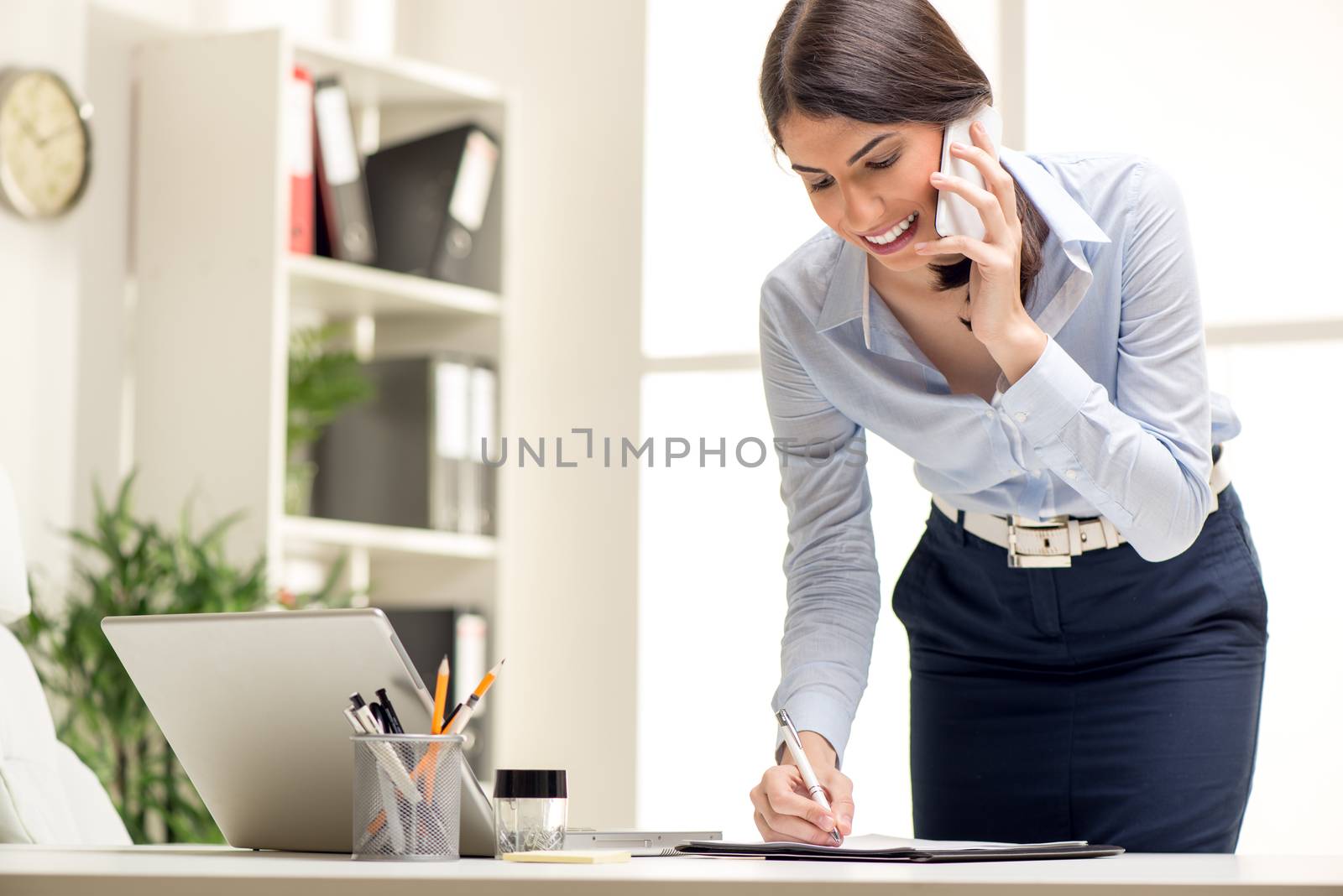 Beautiful Busy Businesswoman, standing in office, writing in planner and talking on mobile phone.