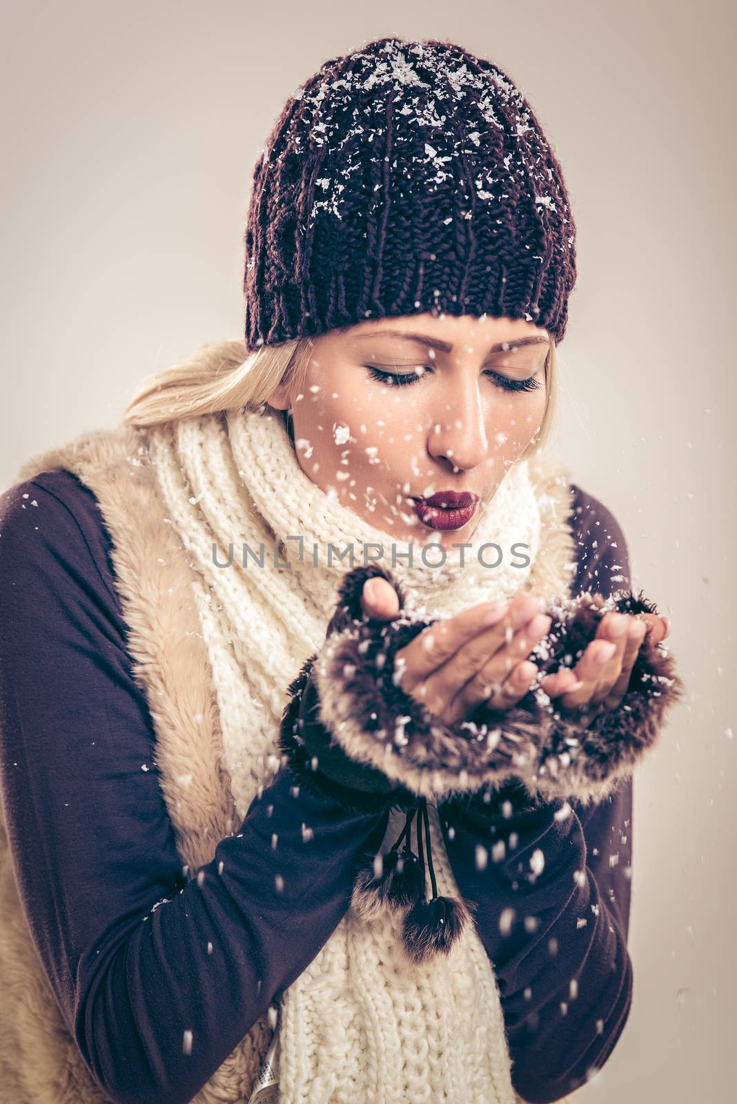Close-up of beautiful young woman in winter clothes as blowing snowflakes which holds in her hands.