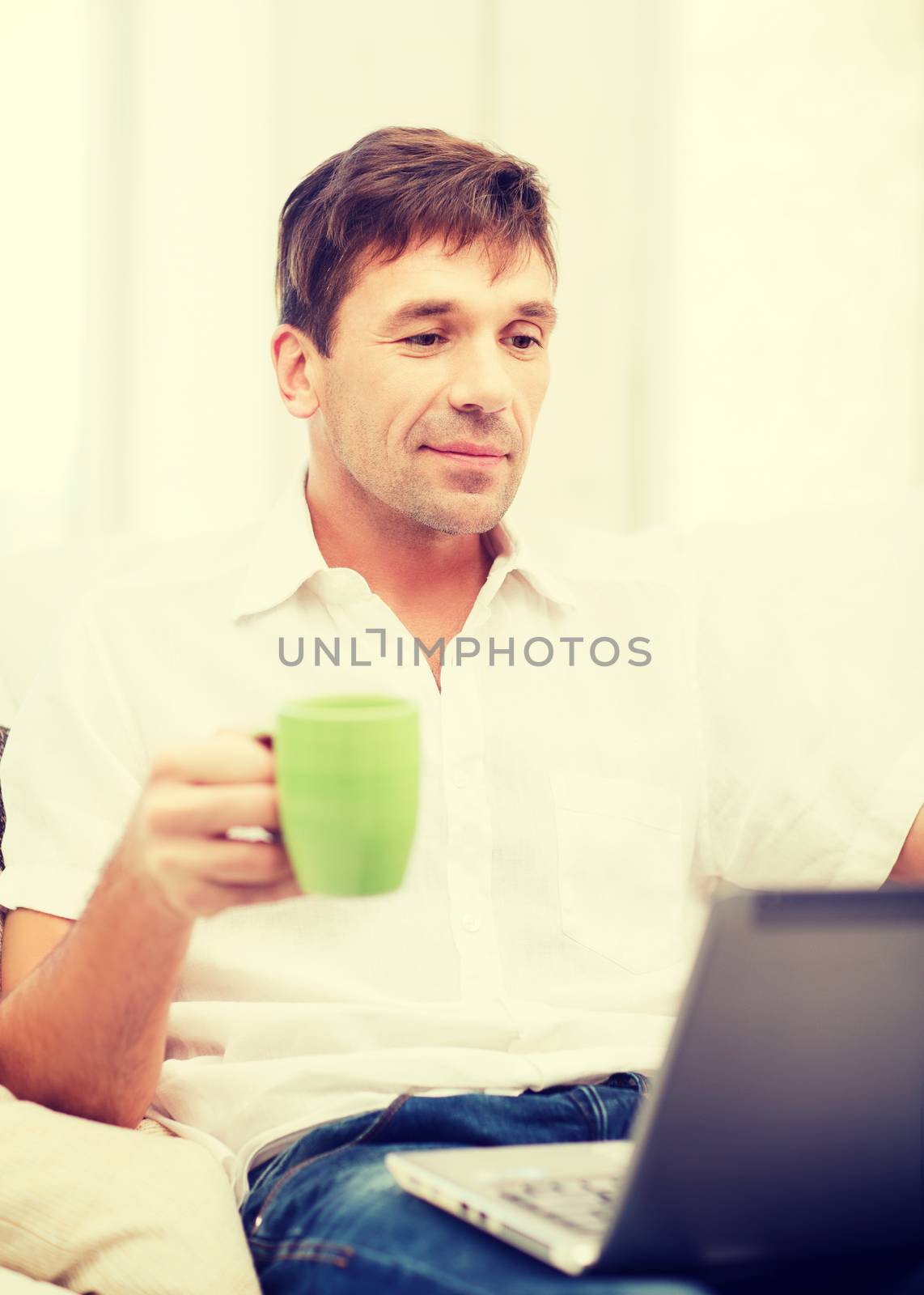 technology, drinks and lifestyle concept - man working with laptop at home, holding a cup of warm tea or coffee