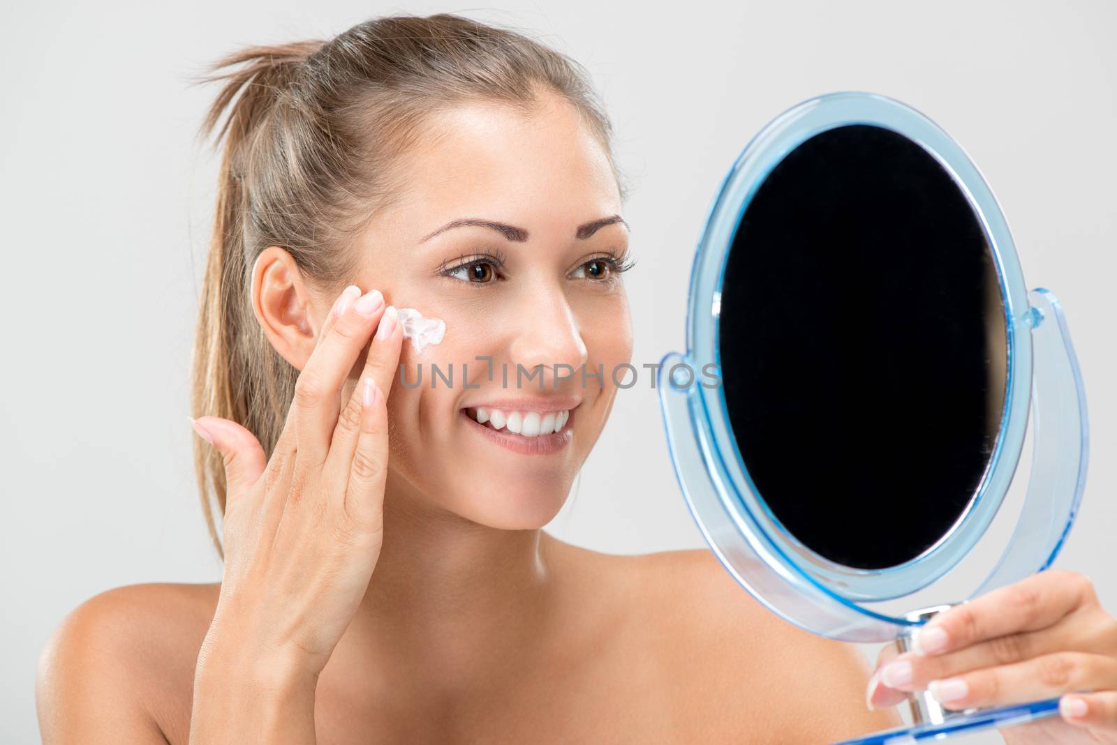 Cute girl preparing to start her day. She is applying moisturizer cream on face. Looking at mirror and smiling. Close-up.