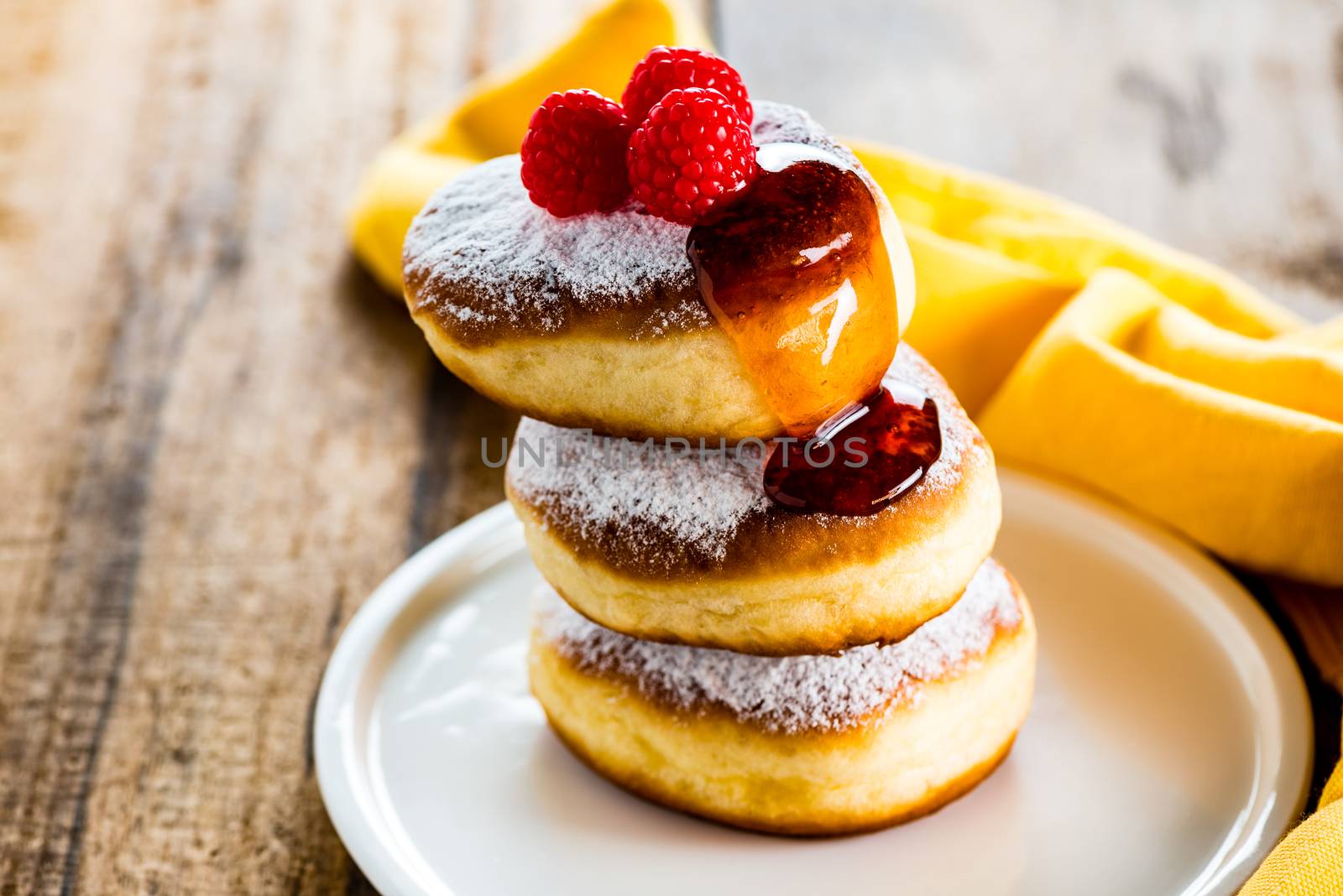 A stack of tree delicious german doughnuts powdered with sugar decorated with raspberry and raspberry sauce on white plate and yellow napkin