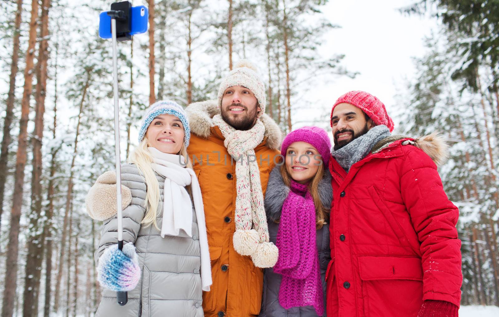 technology, season, friendship and people concept - group of smiling men and women taking selfie with smartphone and monopod in winter forest