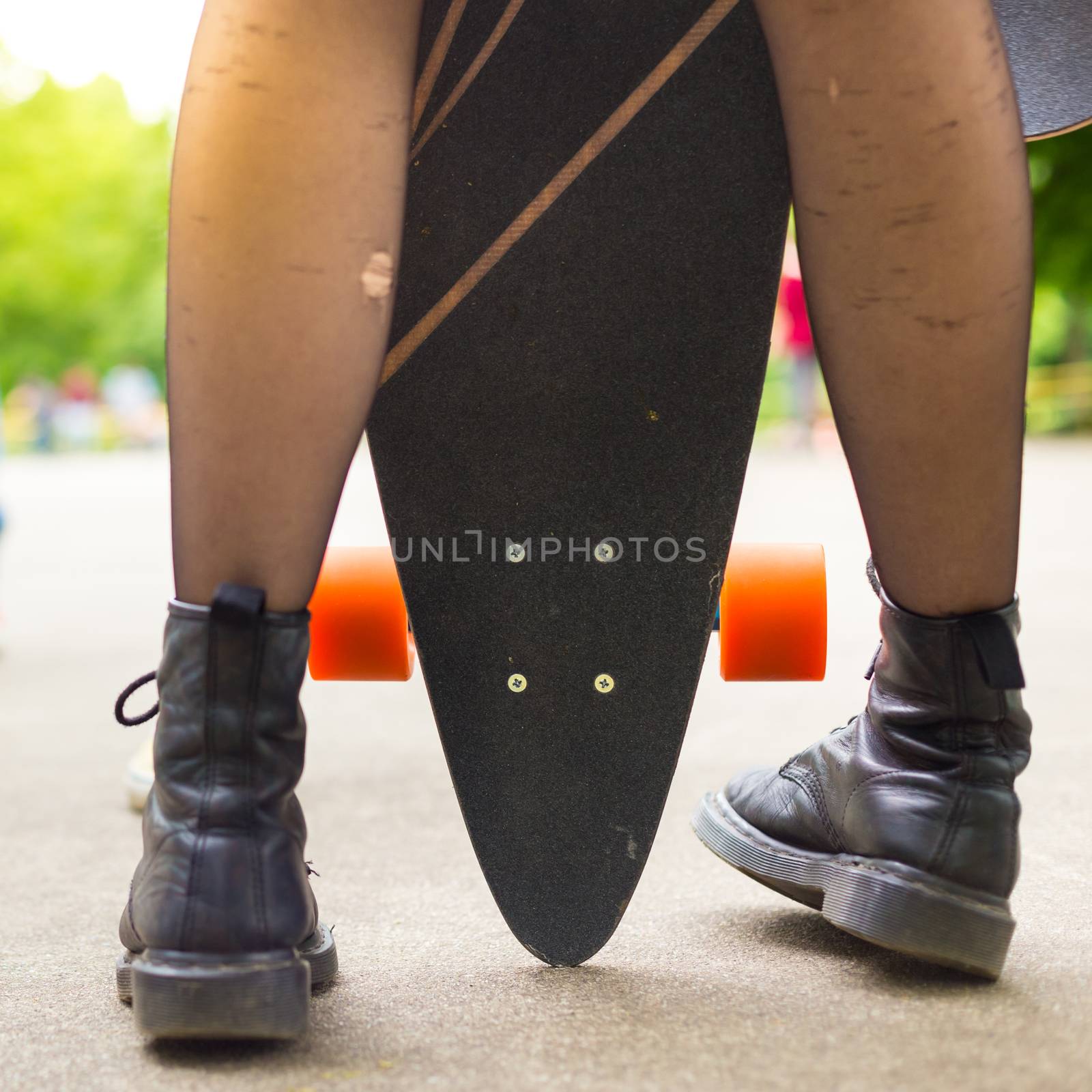 Teenagers practicing long board riding outdoors in skateboarding park. Detail of  legs of girl wearing black boots and stockings holding long board in foreground. Active urban life. Urban subculture.