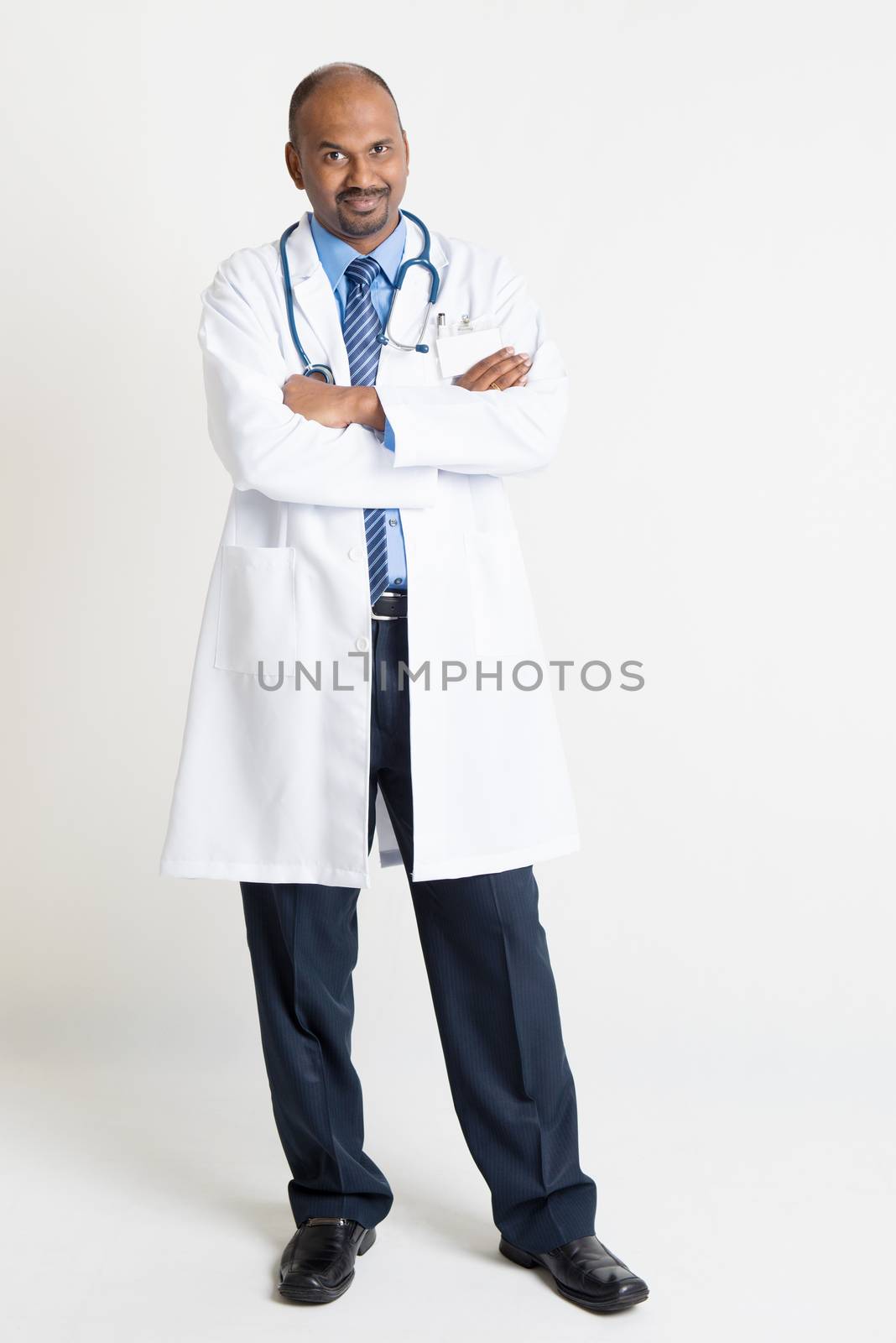 Full length mature Indian male medical doctor in uniform arms crossed standing on plain background with shadow.