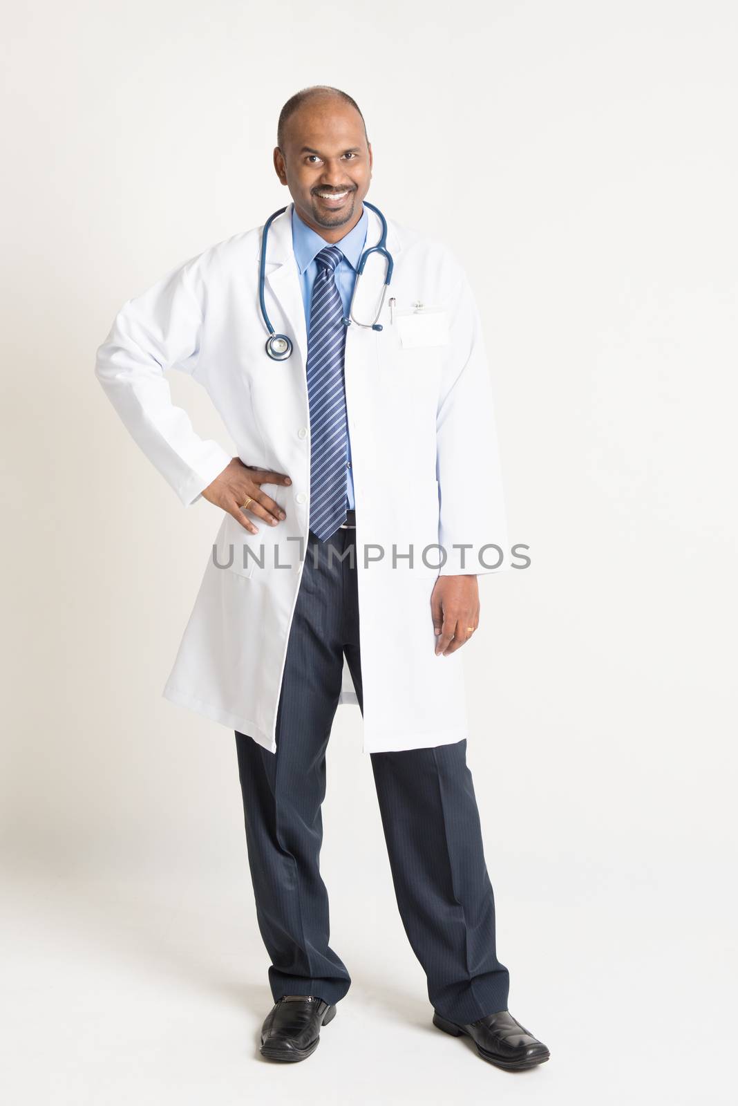 Full length mature Indian male medical doctor in uniform standing on plain background with shadow.