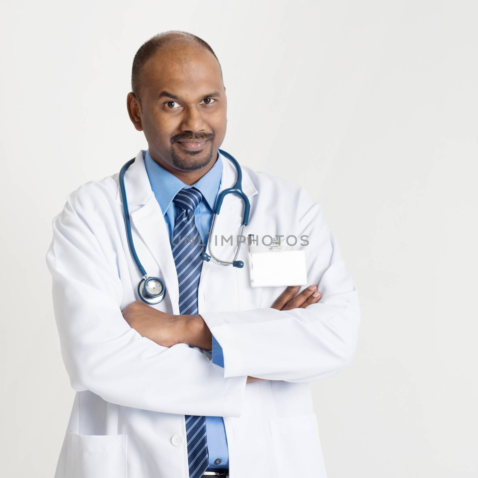 Portrait of mature Indian male medical doctor in uniform standing on plain background with shadow.