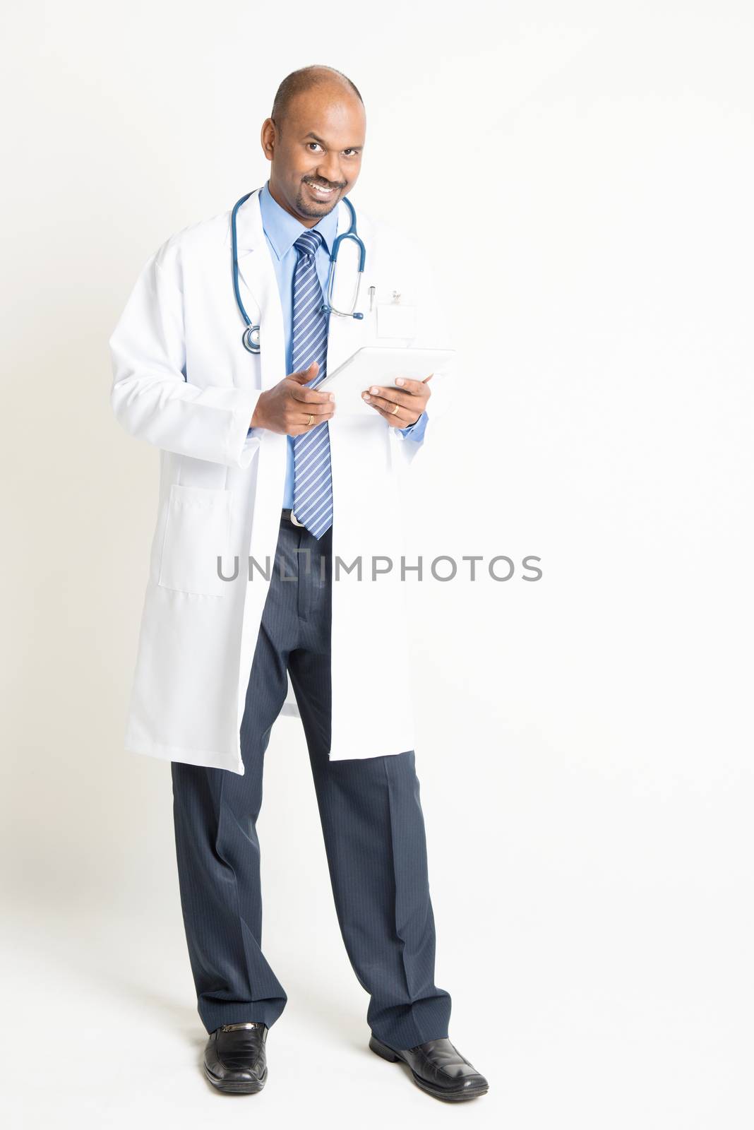 Full length mature Indian male medical doctor in uniform using digital tablet computer, standing on plain background with shadow.
