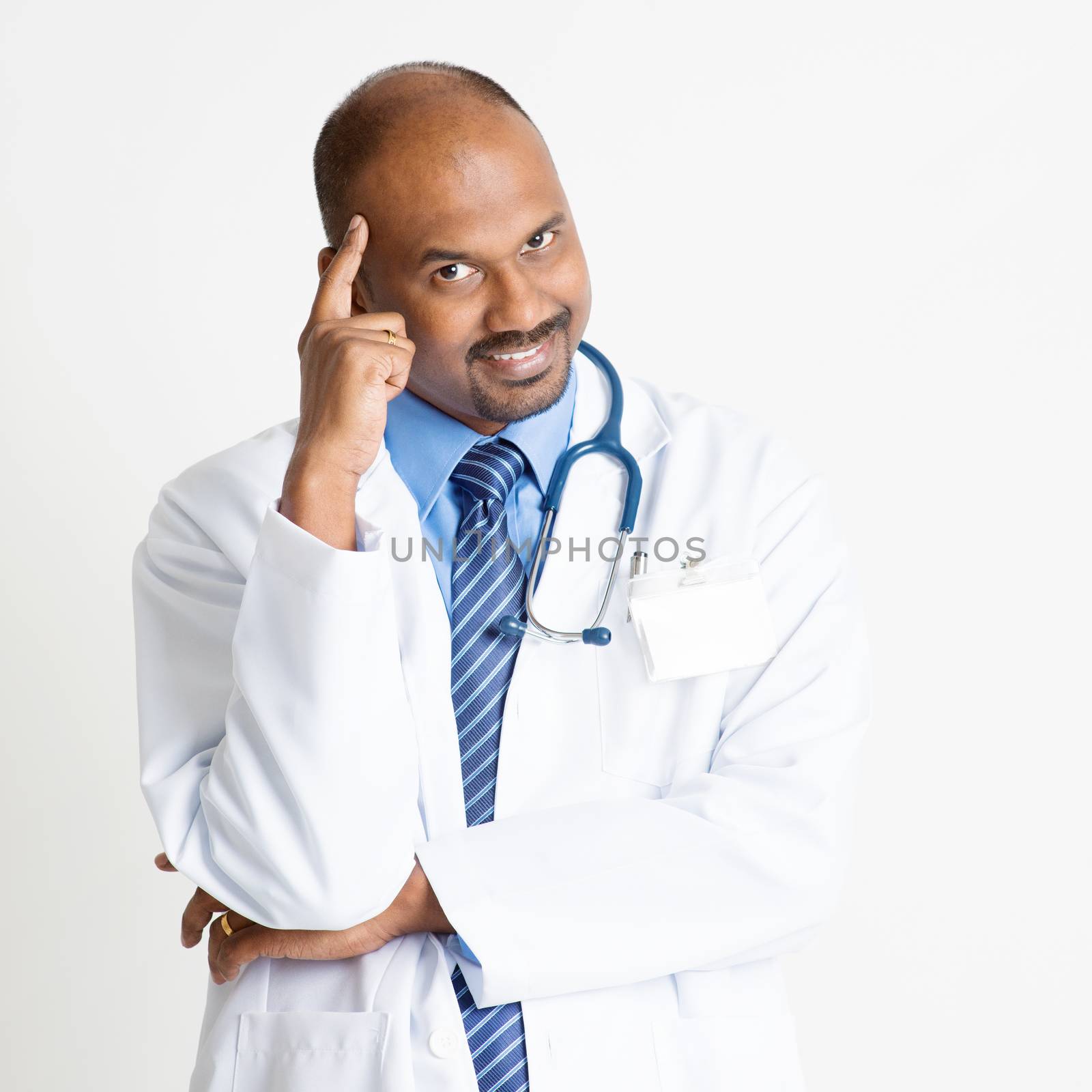 Portrait of mature Indian male medical doctor in uniform thinking, standing on plain background with shadow.