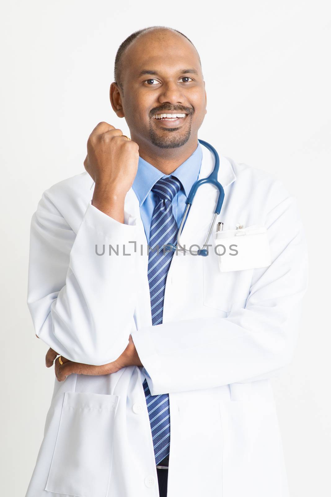 Portrait of happy mature Indian male medical doctor in uniform smiling, standing on plain background with shadow.