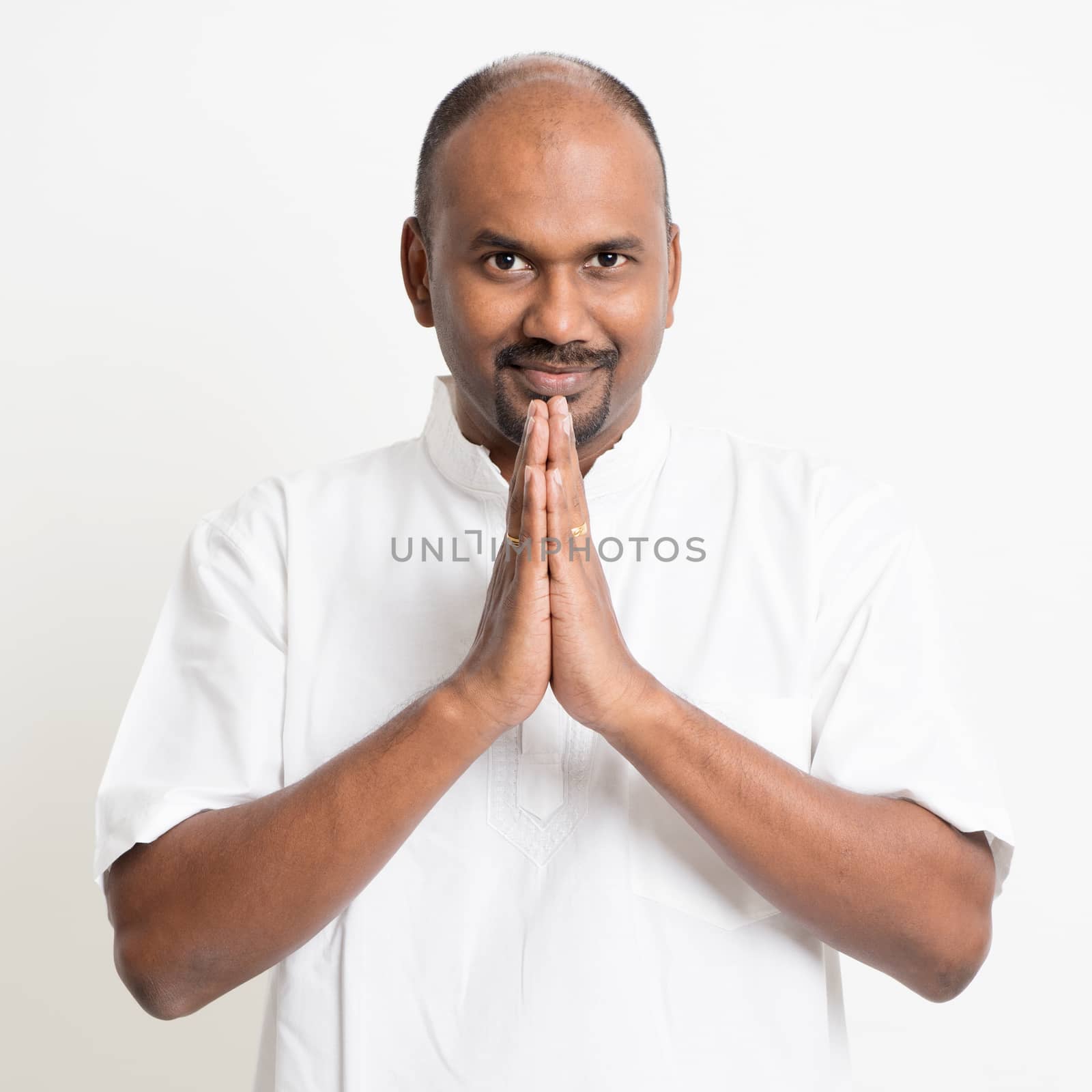Portrait of mature casual business Indian man praying, standing on plain background with shadow.