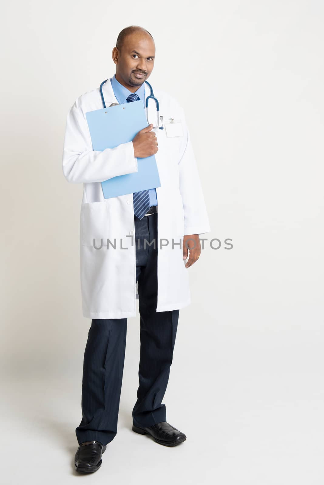 Full length mature Indian male medical doctor in uniform holding medical report folder, standing on plain background with shadow.