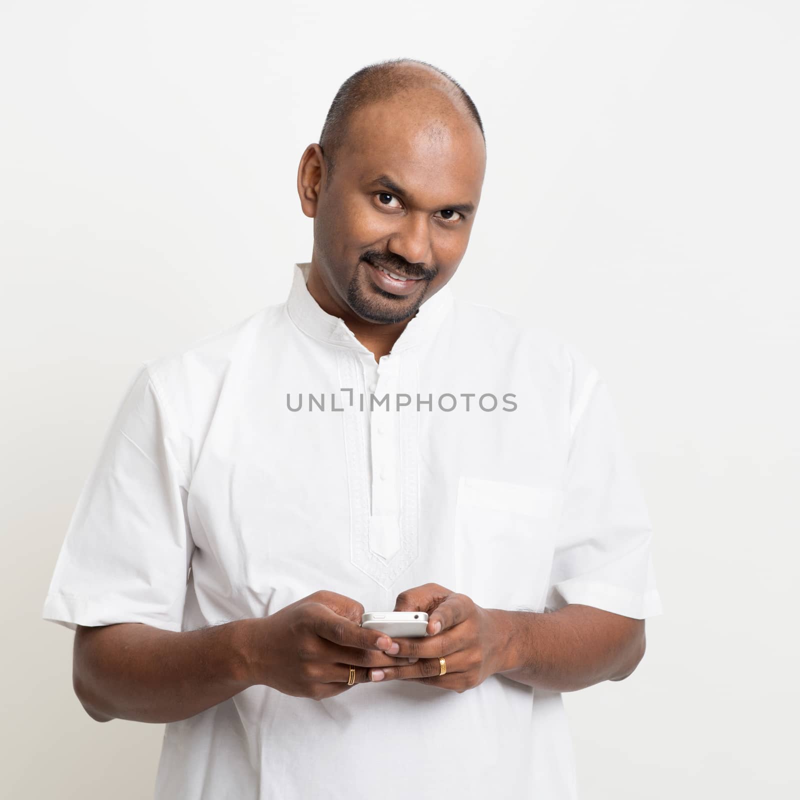 Portrait of mature casual business Indian man texting using smartphone, standing on plain background with shadow.