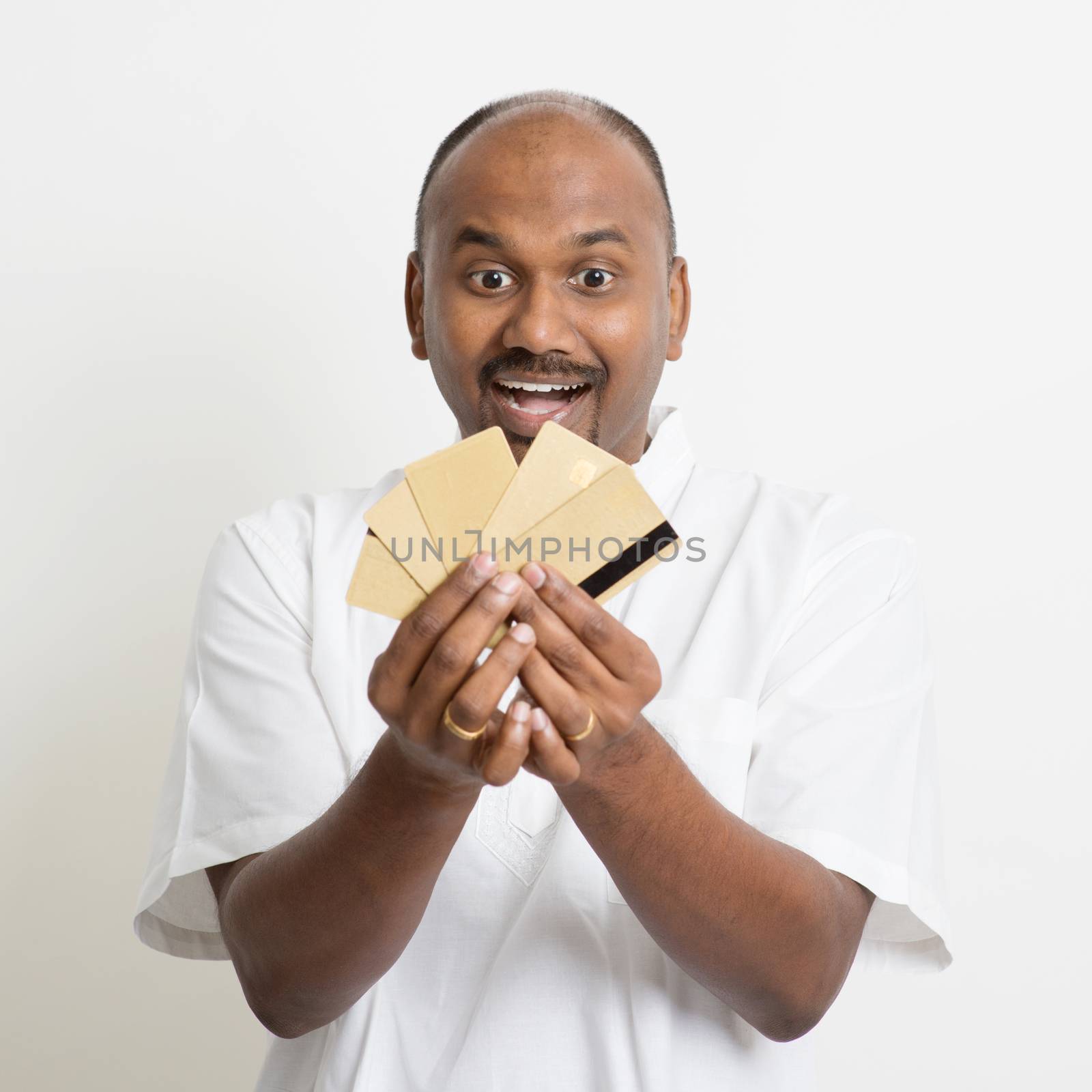 Portrait of mature casual business Indian man holding many credit cards and laughing, standing on plain background with shadow.