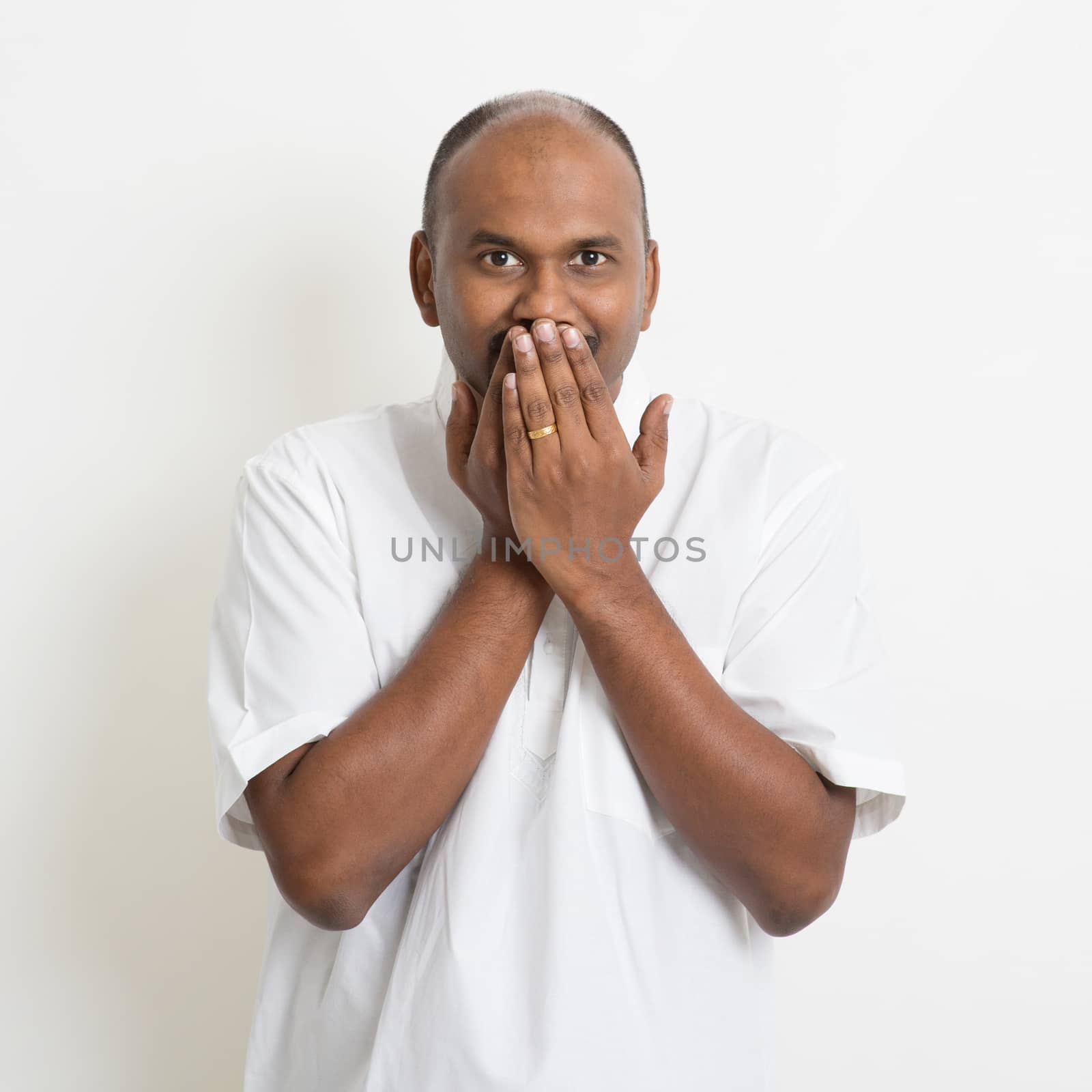 Portrait of mature casual business Indian man covered mouth, standing on plain background with shadow.