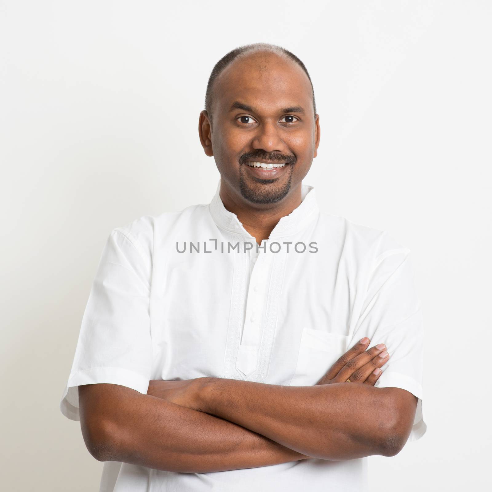 Portrait of mature casual business Indian man arms crossed and smiling, standing on plain background with shadow.