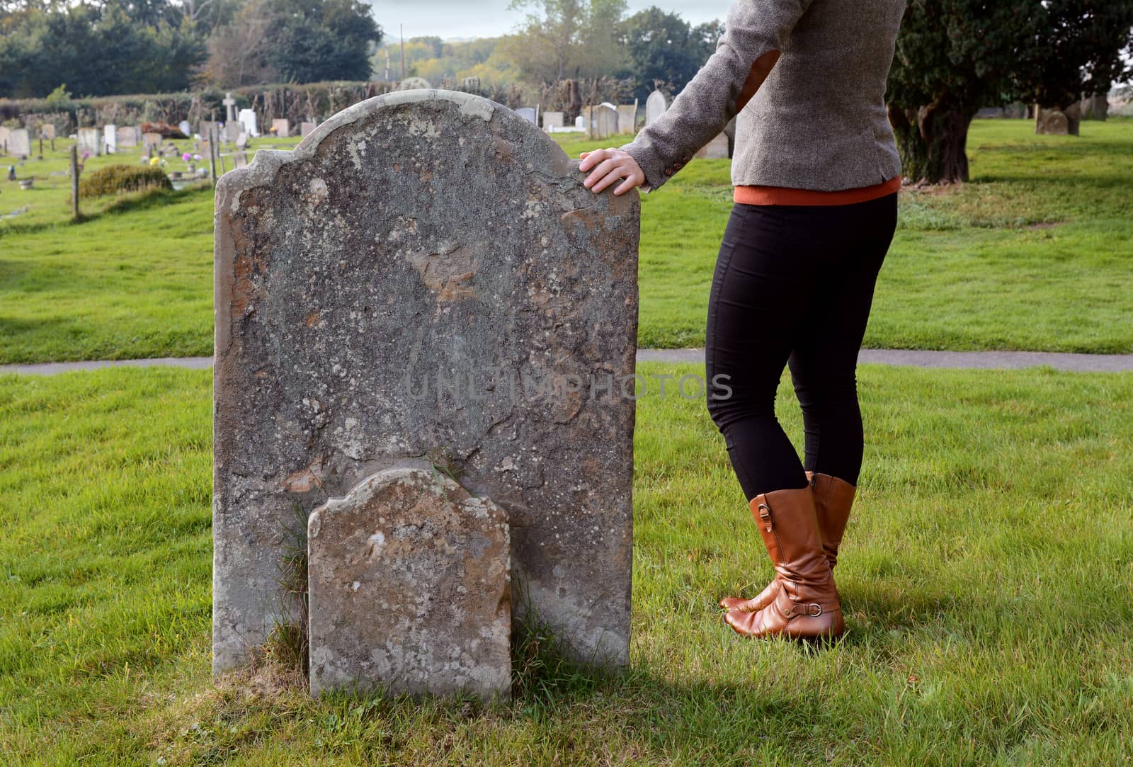 Smartly dressed woman rests her hand thoughtfully on a gravestone in a cemetery