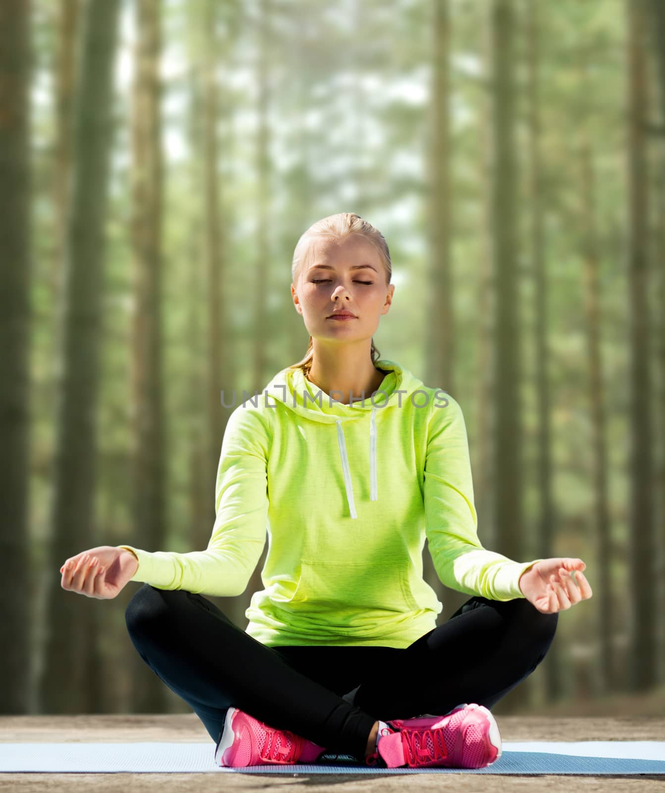 sport, fitness, yoga and people concept - happy young woman meditating in lotus pose and sitting on mat over woods background