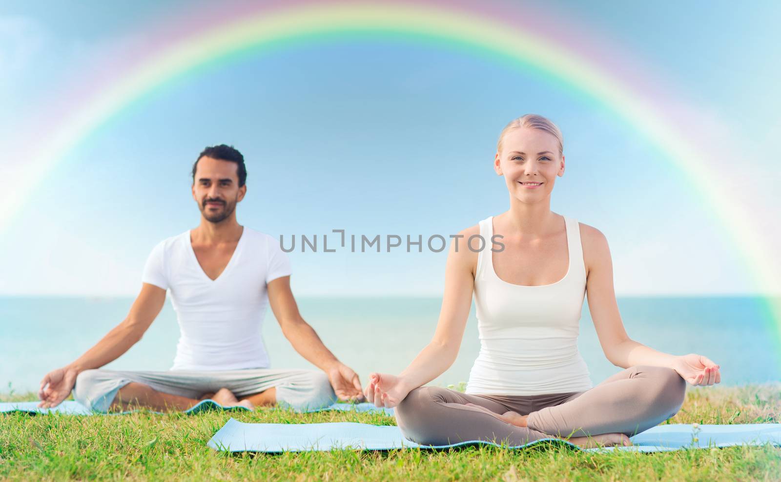 sport, fitness, yoga and people concept - smiling couple meditating and sitting on mats over sea and rainbow in blue sky background