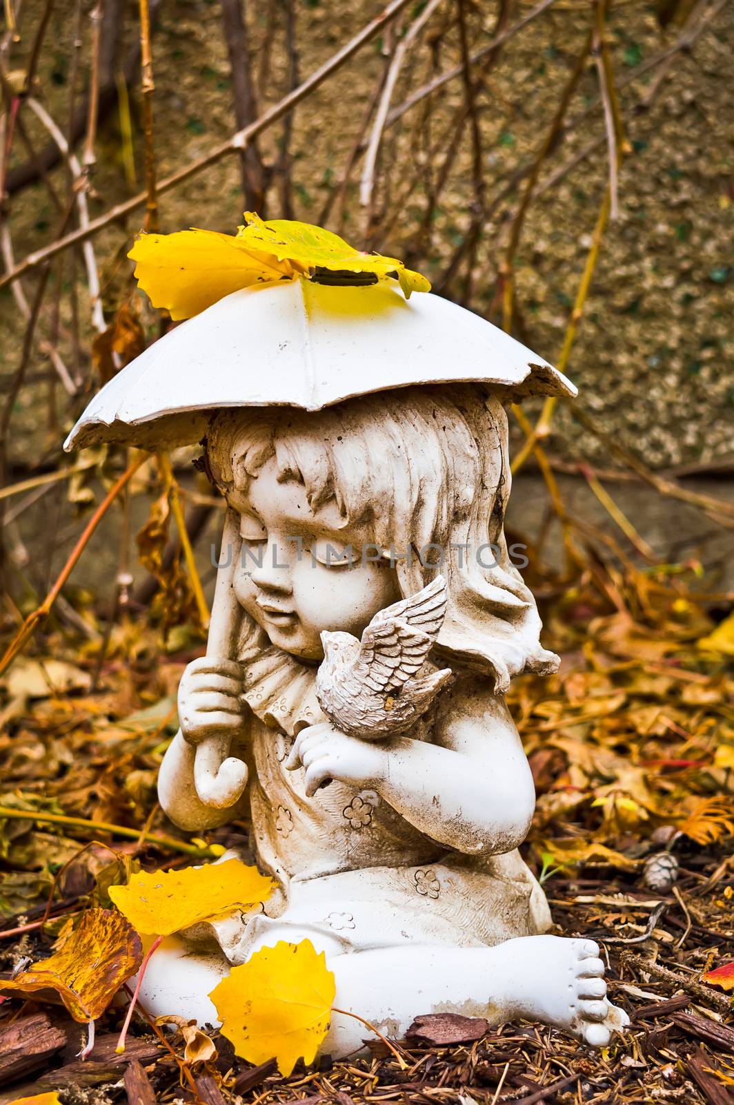 Profile of a small weathered statue of a young girl holding an umbrella and a bird, surrounded by dry leaves and bark chips in my backyard.