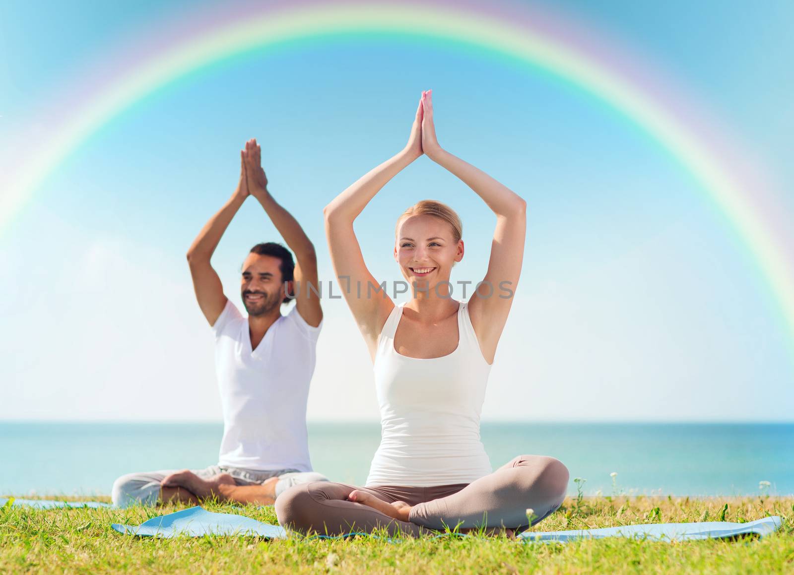sport, fitness, yoga and people concept - smiling couple meditating and sitting on mats with raised hands over sea and rainbow in blue sky background