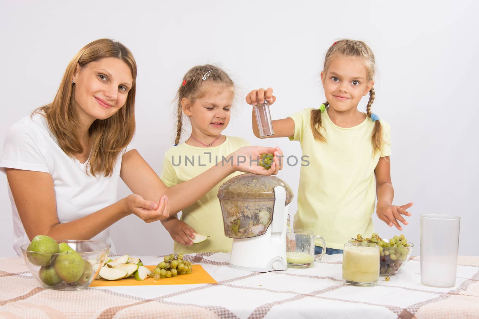 Young beautiful mother and two daughters sitting at a table squeezed juice from pears and grapes with a juicer