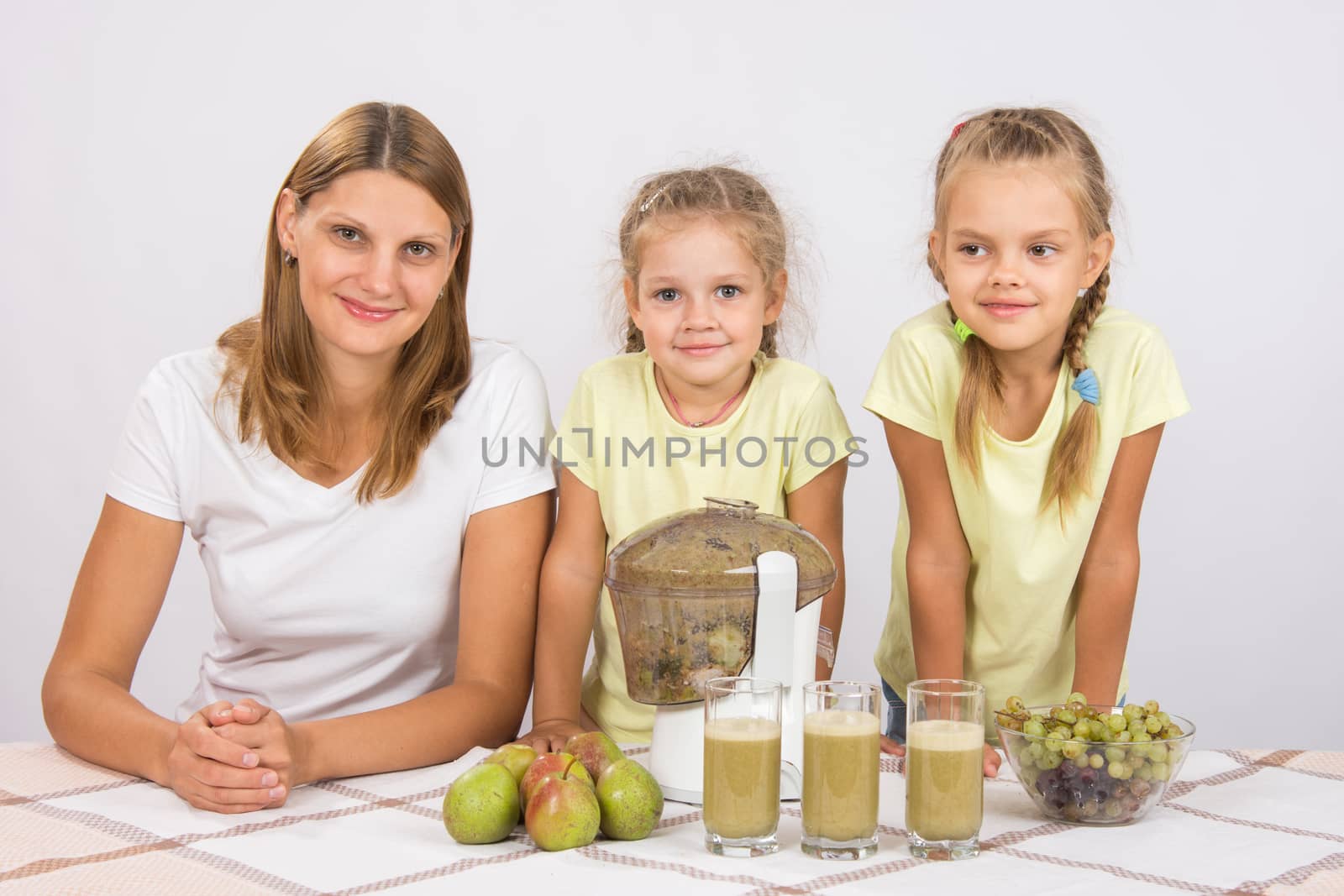 Young beautiful mother and two daughters sitting at a table squeezed juice from pears and grapes with a juicer