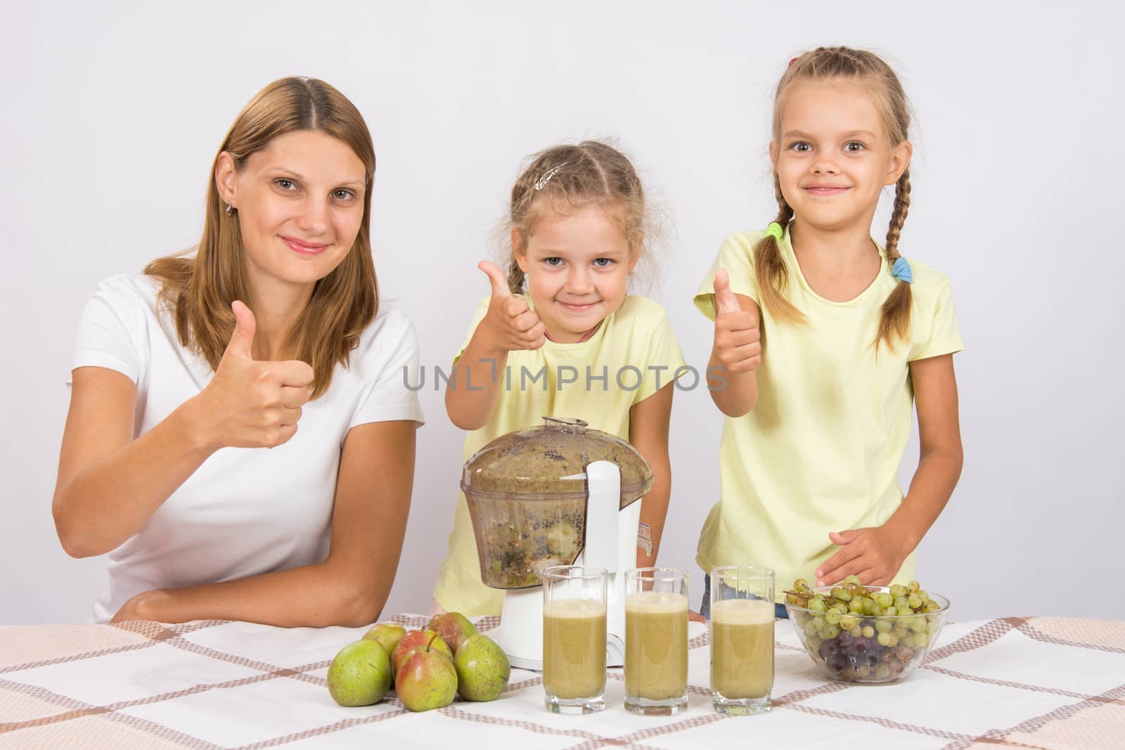 Young beautiful mother and two daughters sitting at a table squeezed juice from pears and grapes with a juicer