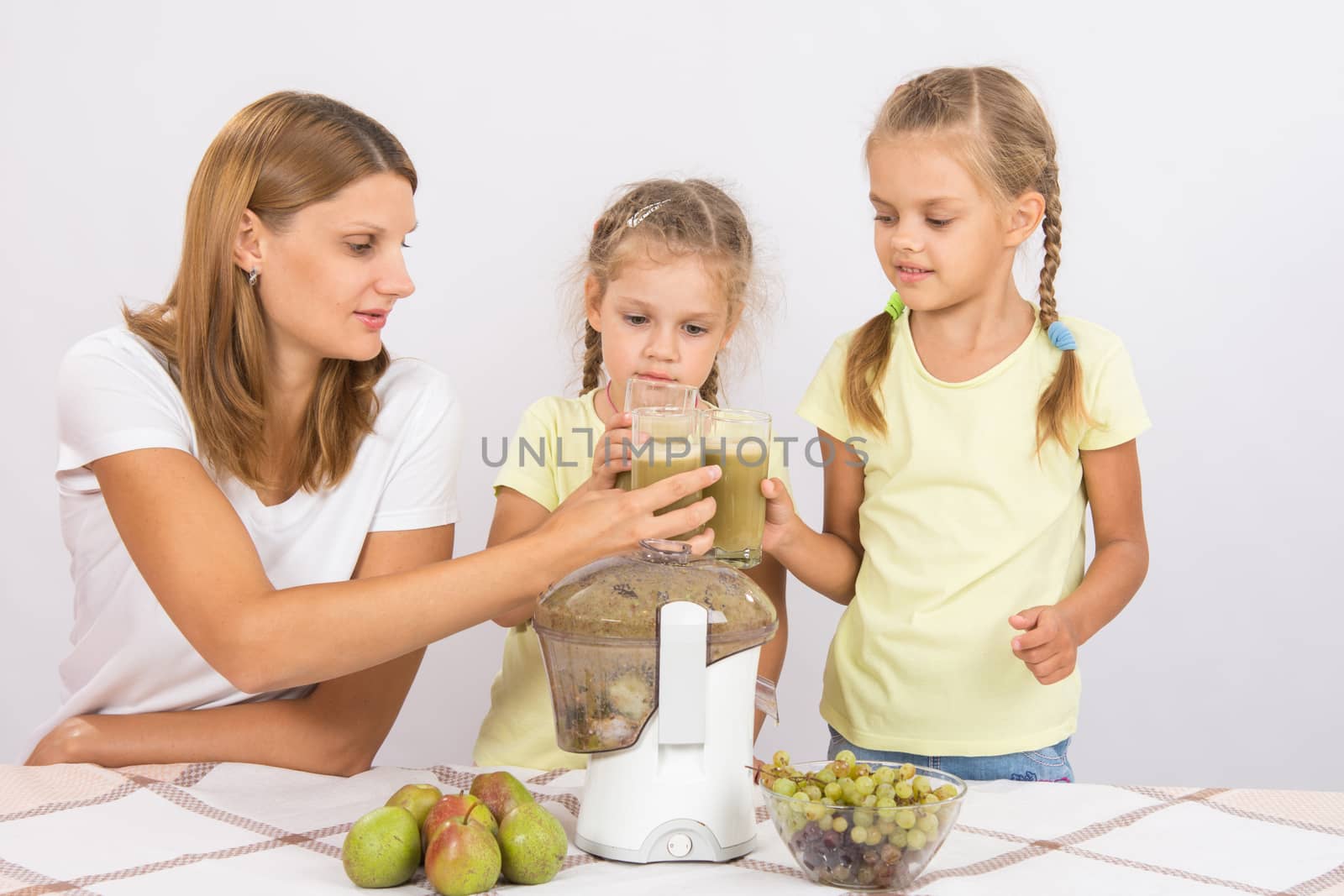 Young beautiful mother and two daughters sitting at a table squeezed juice from pears and grapes with a juicer
