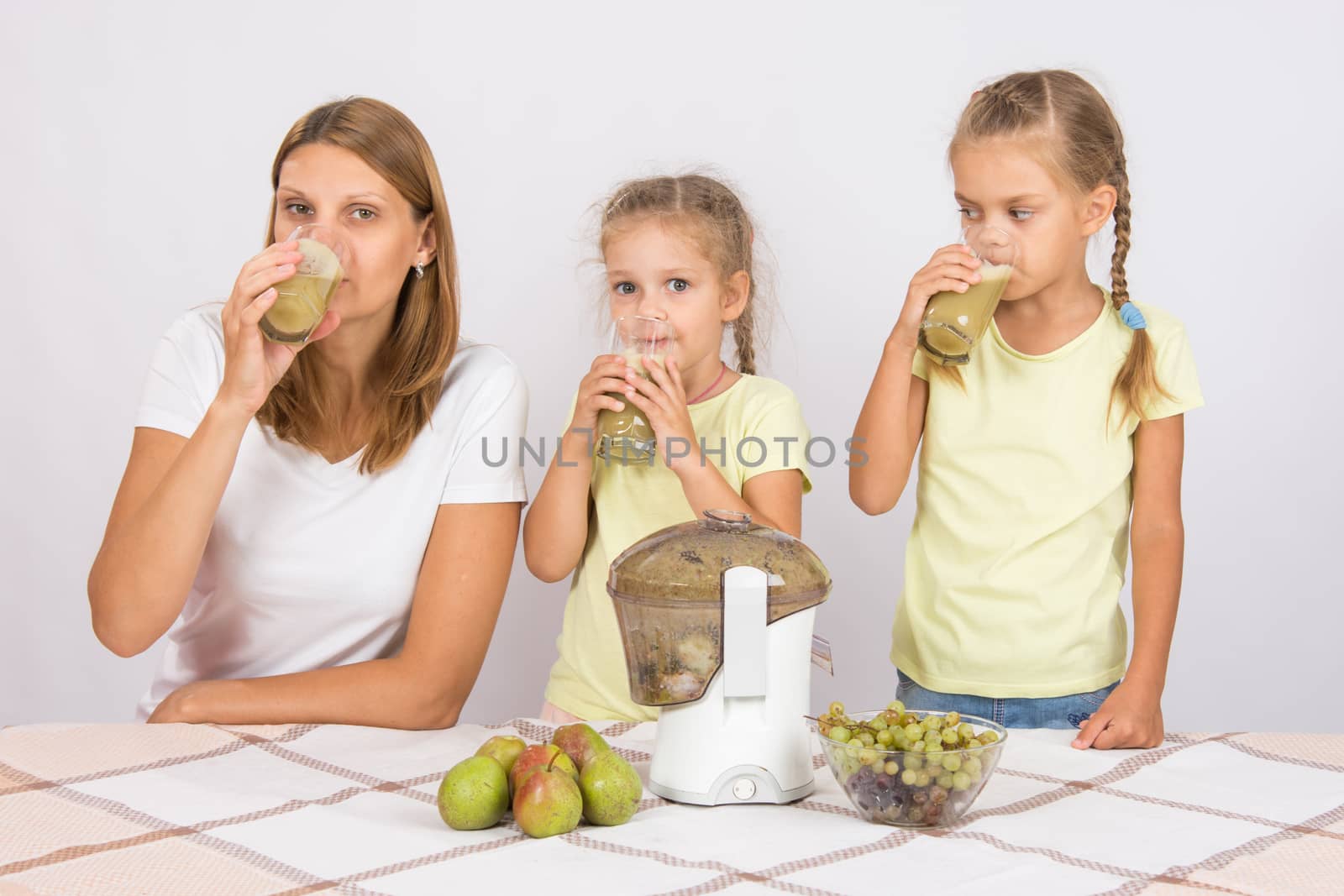 Young beautiful mother and two daughters sitting at a table squeezed juice from pears and grapes with a juicer