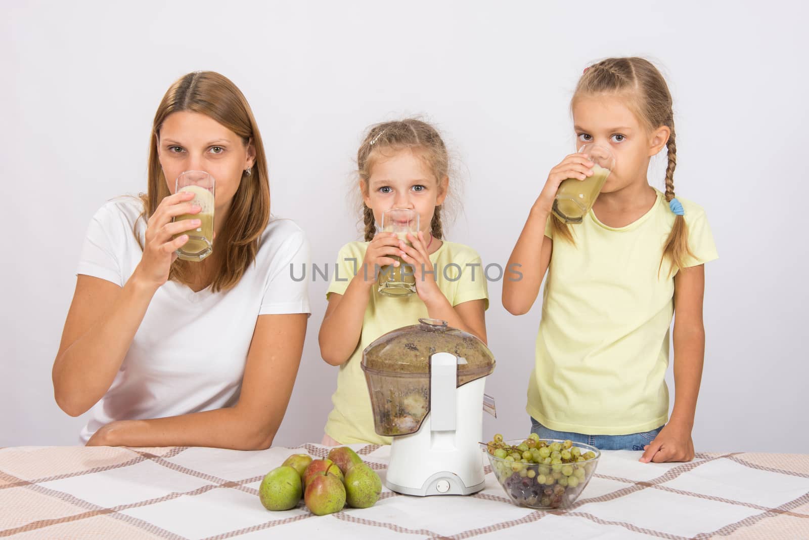 Young beautiful mother and two daughters sitting at a table squeezed juice from pears and grapes with a juicer