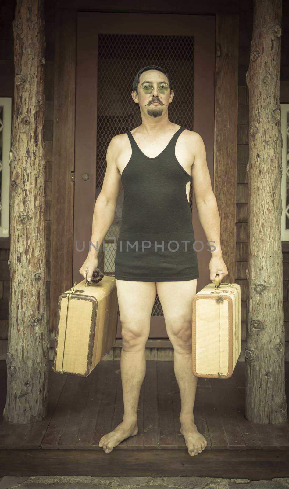 Goofy Gentleman Dressed in 1920’s Era Swimsuit Holding Suitcases on Porch of Cabin.