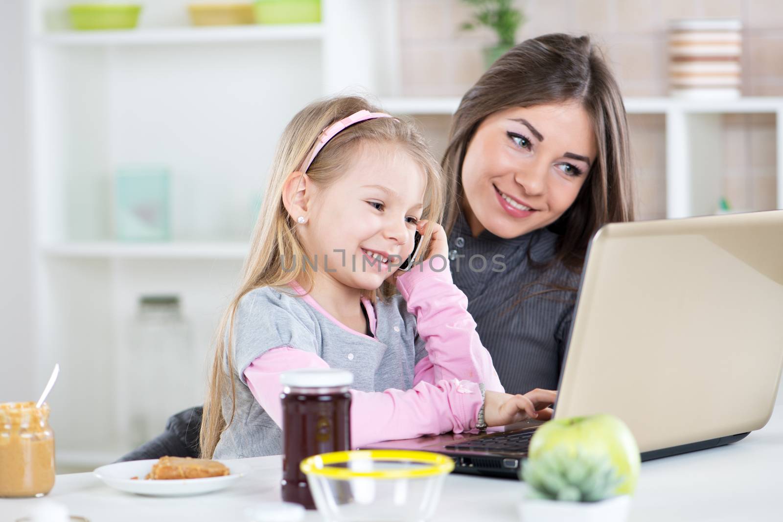 Mom and daughter having fun in the morning during breakfast. Cute Little girl imitates mom to use laptop and telephone.